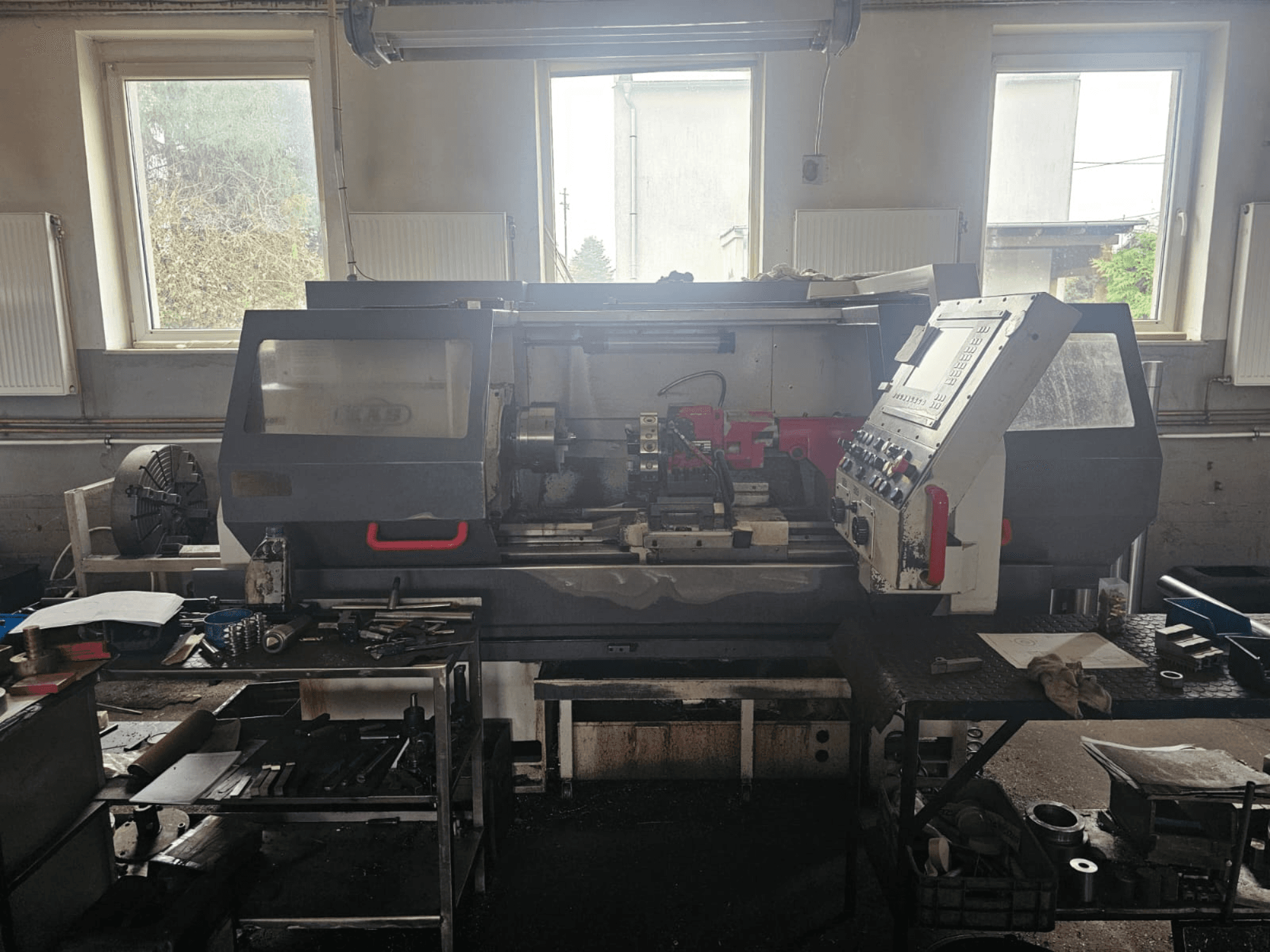 Draaibankmachine in een werkplaats, vooraanzicht, met bedieningspaneel en gereedschappen op tafels, omgeven door ramen en een rommelige werkruimte.