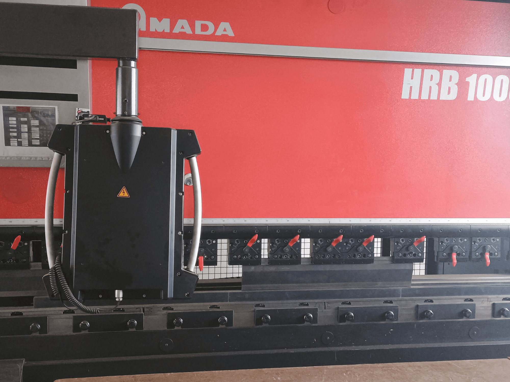 Amada HRB 100 buigmachine vooraanzicht; heeft een zwarte besturingskop met een conisch mondstuk en verstelbare klemmen.