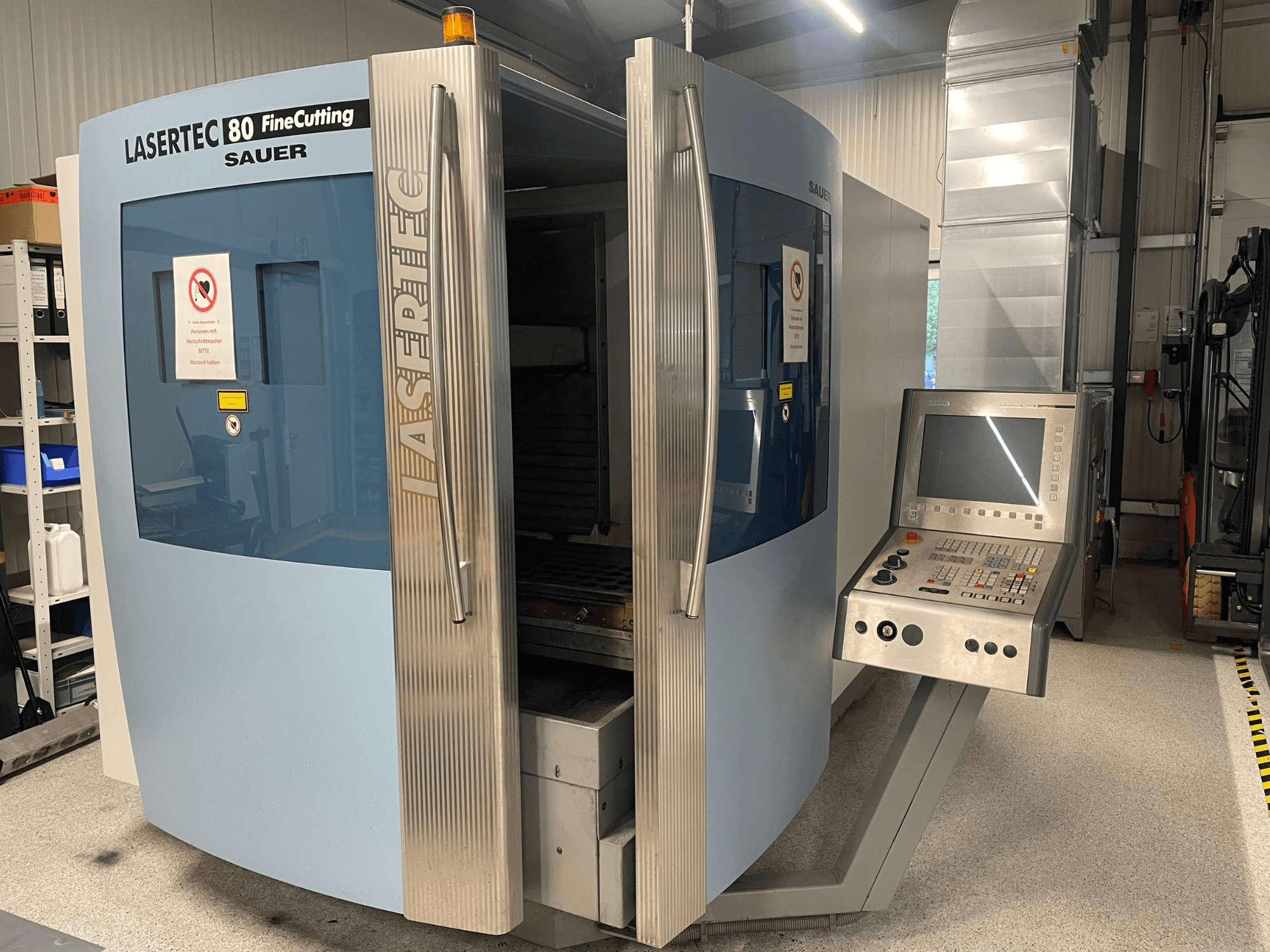 Vooraanzicht van de DMG Sauer Lasertec80 FineCutting-plaatbewerkingsmachine met bedieningspaneel, open deuren en zichtbare veiligheidsborden.