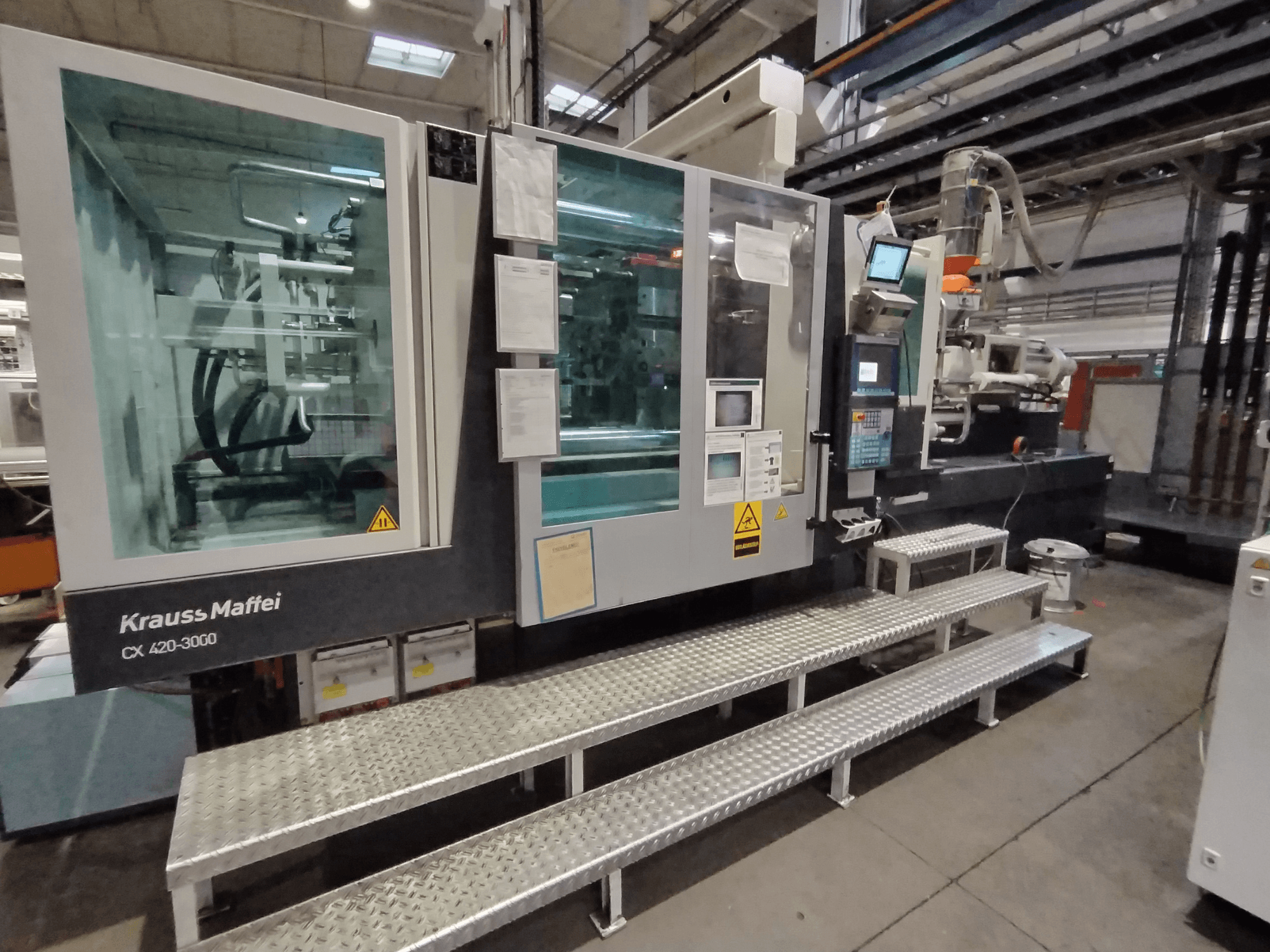 Krauss Maffei CX 420-3000 spuitgietmachine, zijaanzicht, met bedieningspaneel en veiligheidssignalering zichtbaar, opgesteld op metalen treden.