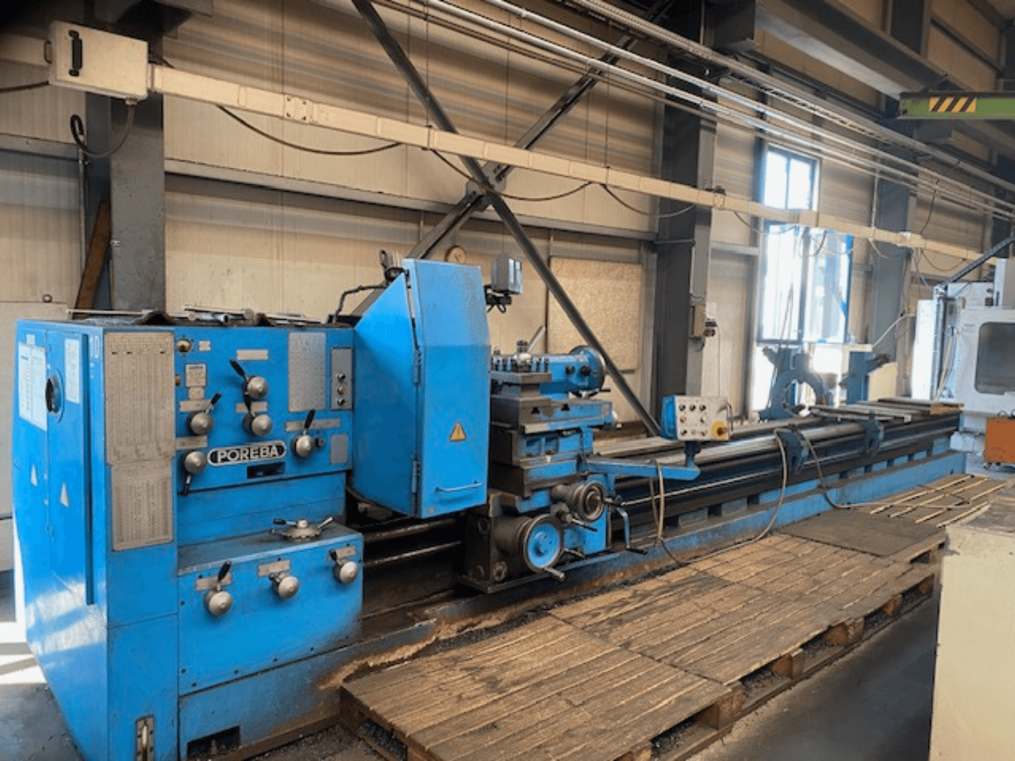 Vooraanzicht van een blauwe POREBA draaibank, met bedieningsknoppen, een gereedschapsteun en houten palletten eronder.