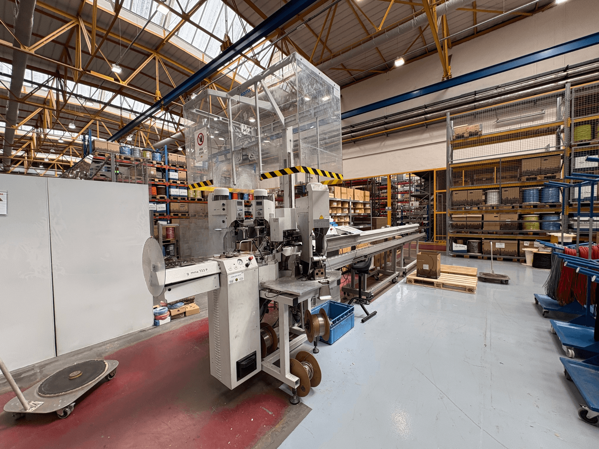 KOMAX GAMMA 333 PC bewerkingsmachine in een magazijn, zijaanzicht met bedieningselementen, transportsysteem en omringende schappen.