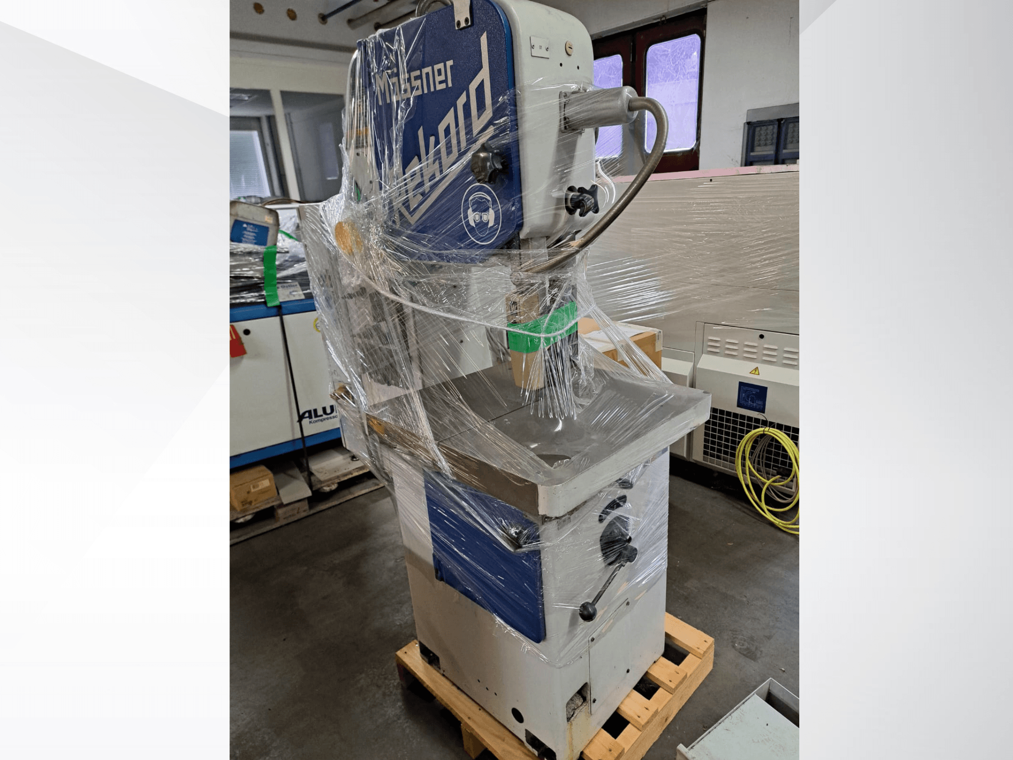 MOSSNER Rekord metalen lintzaag, verpakt in plastic; zijaanzicht met bedieningspaneel en onderstel op een houten pallet in een werkplaats.