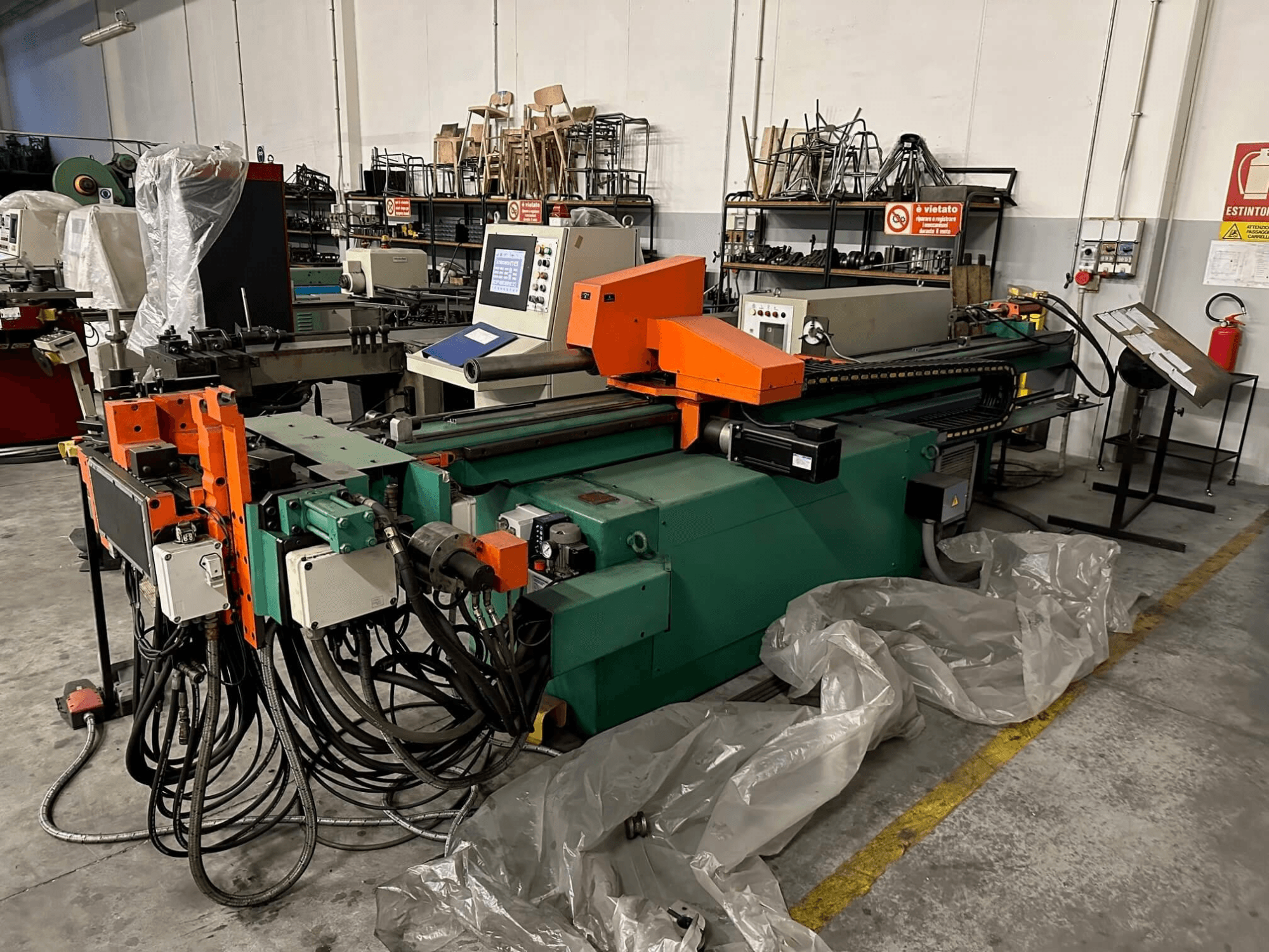 Industriële machine met een oranje en groene behuizing, bedieningspaneel en talloze kabels, gezien vanaf de voorkant in een werkplaats.