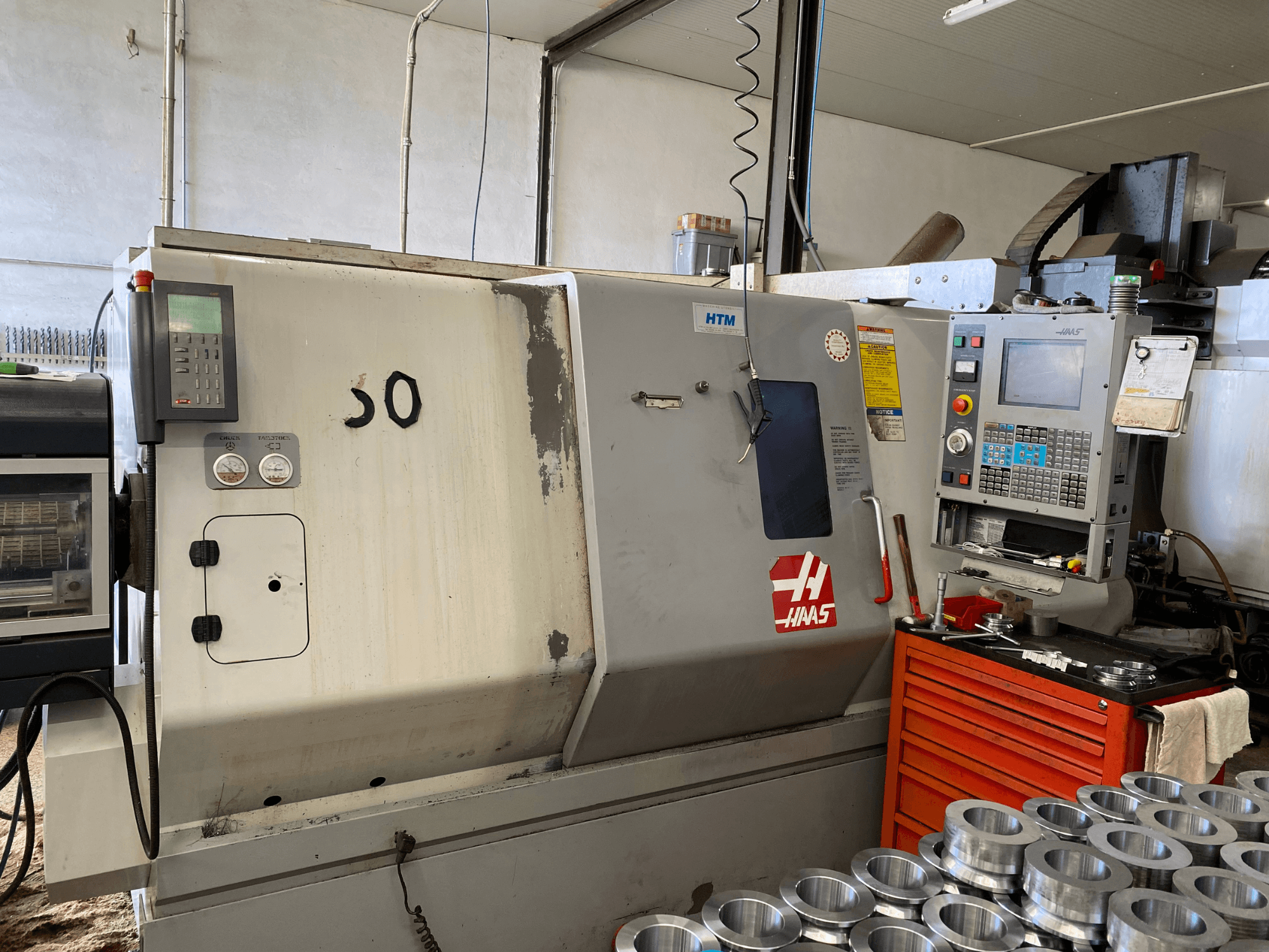 Haas CNC machine met bedieningspaneel, vooraanzicht, met bedieningsknoppen, meters en gestapelde metalen onderdelen in de buurt.