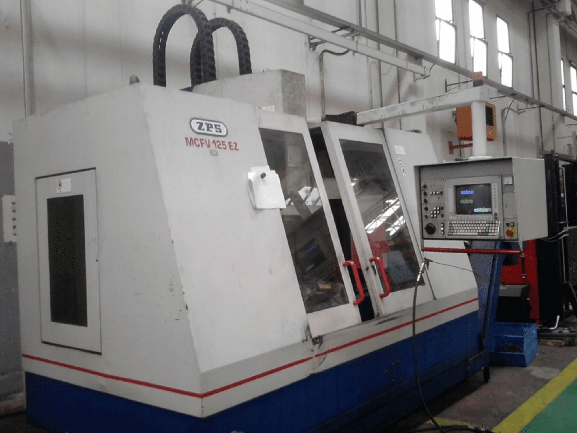 ZPS MCFV 125 EZ CNC machine, vooraanzicht met bedieningspaneel en rode hendels, in een werkplaats met zichtbare slangen.