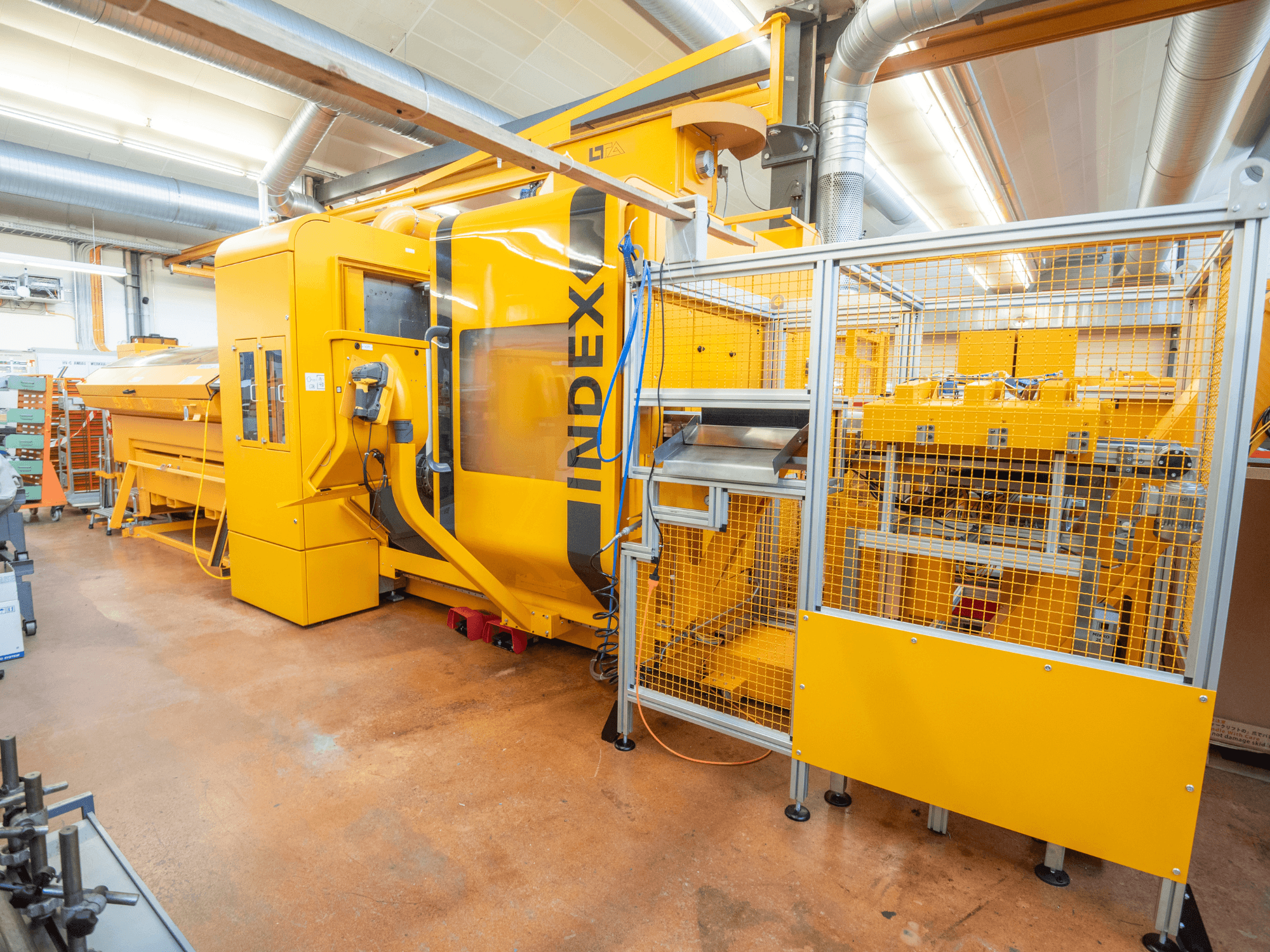 Gele INDEX-machine in een werkplaats, vooraanzicht. Met veiligheidsbehuizing, bedieningspaneel en meerdere operationele onderdelen.