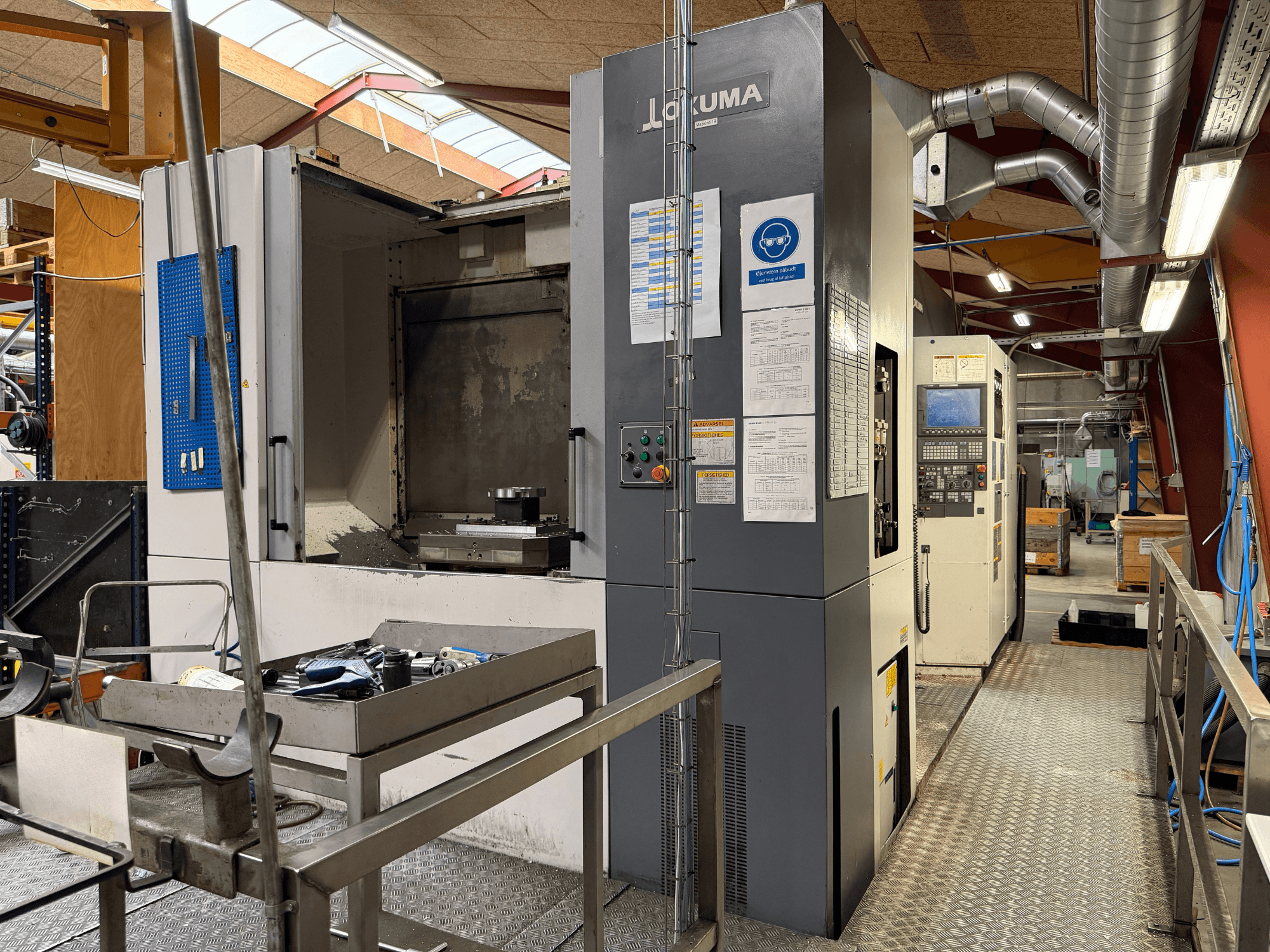 Okuma MA-600HB horizontaal bewerkingscentrum, vooraanzicht, met gereedschappen op een metalen tafel en de bedieningselementen van de machine.