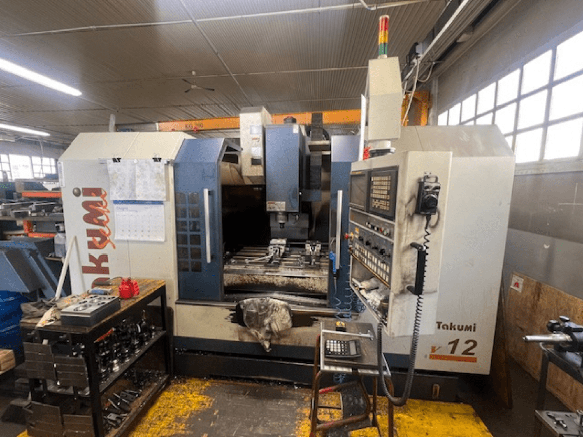 Takumi V12 CNC freesmachine in zijaanzicht, met bedieningspaneel, snijgedeelte en gereedschappen op het werkstation.