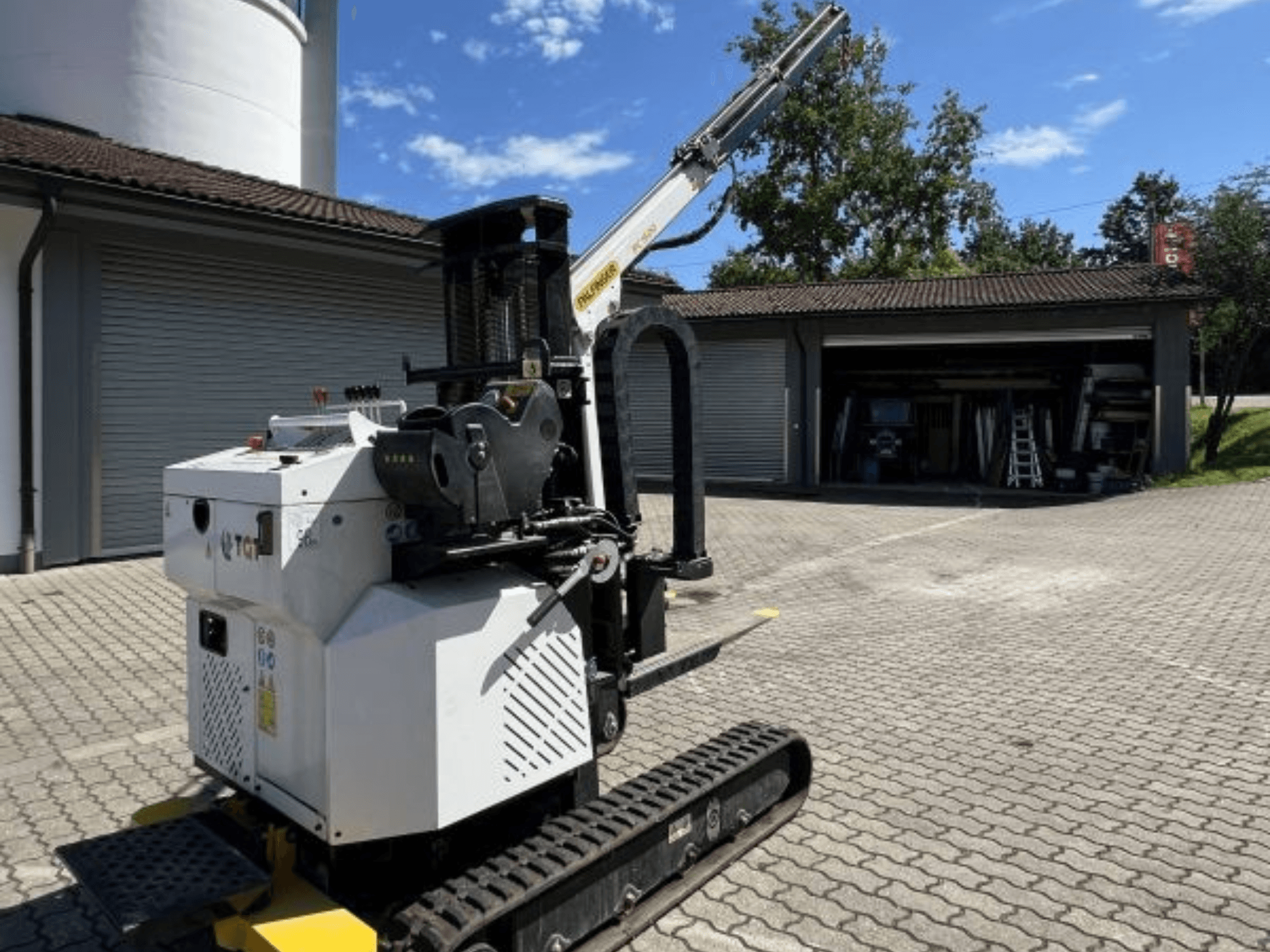 TGI bouwminikraan, zijaanzicht, met rupsen en gemonteerde giek, geparkeerd op een stenen ondergrond bij een garage.