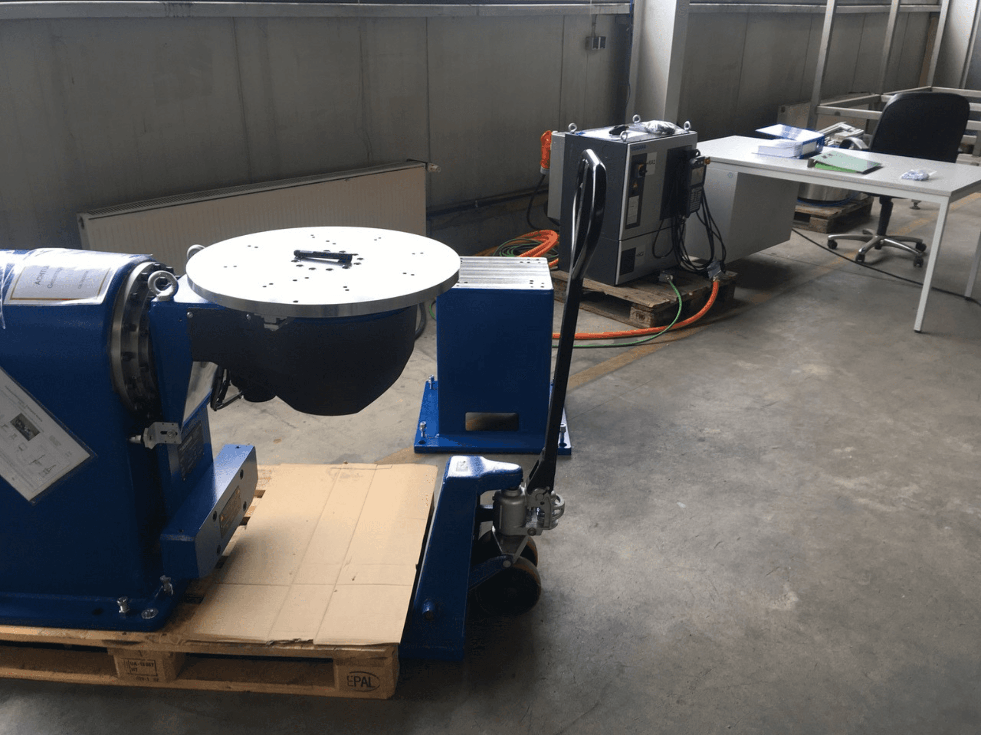 Blauwe industriële apparatuur met een ronde basis en bedieningspaneel, gezien vanaf de voorkant, op een houten pallet in een kantooromgeving.