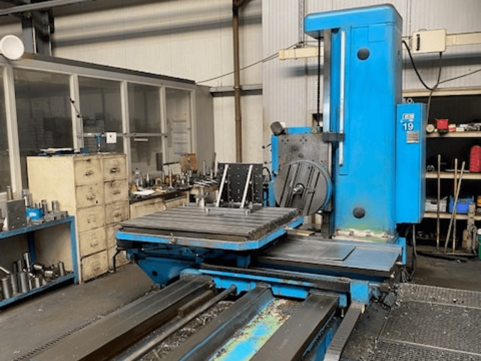 Blauwe freesmachine in vooraanzicht, modelnummer 19, op een werkvloer met gereedschap en een opbergkast op de achtergrond.