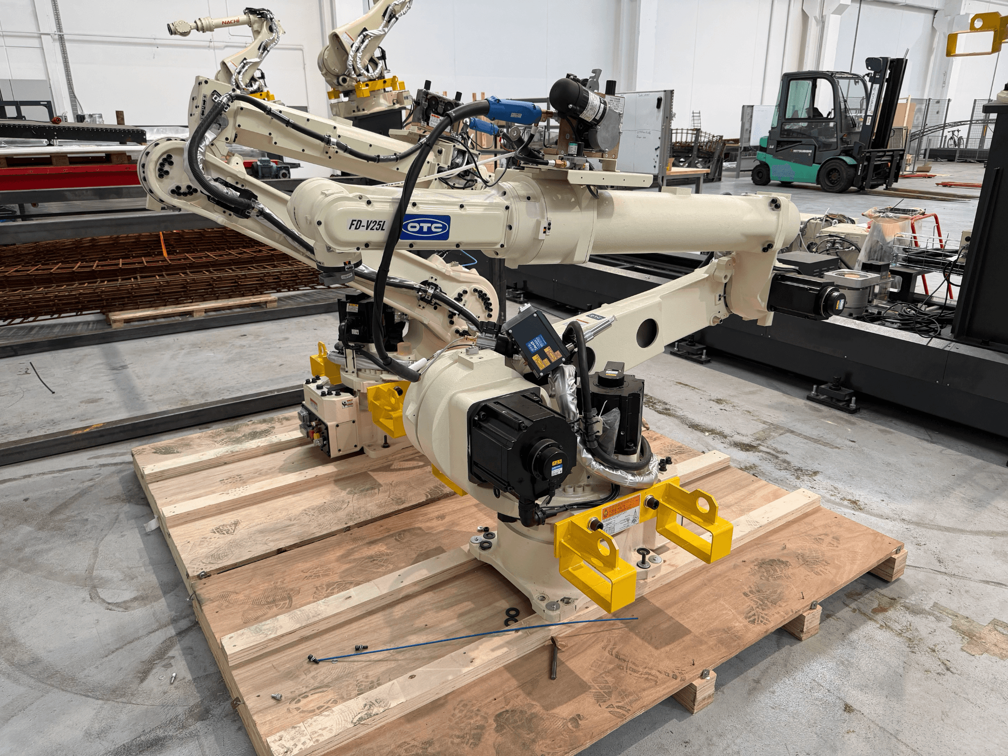 OTC Daihen FD19-V25L-robotarm in vooraanzicht, met gele grijpklauwen en zichtbare mechanische onderdelen op een houten pallet.