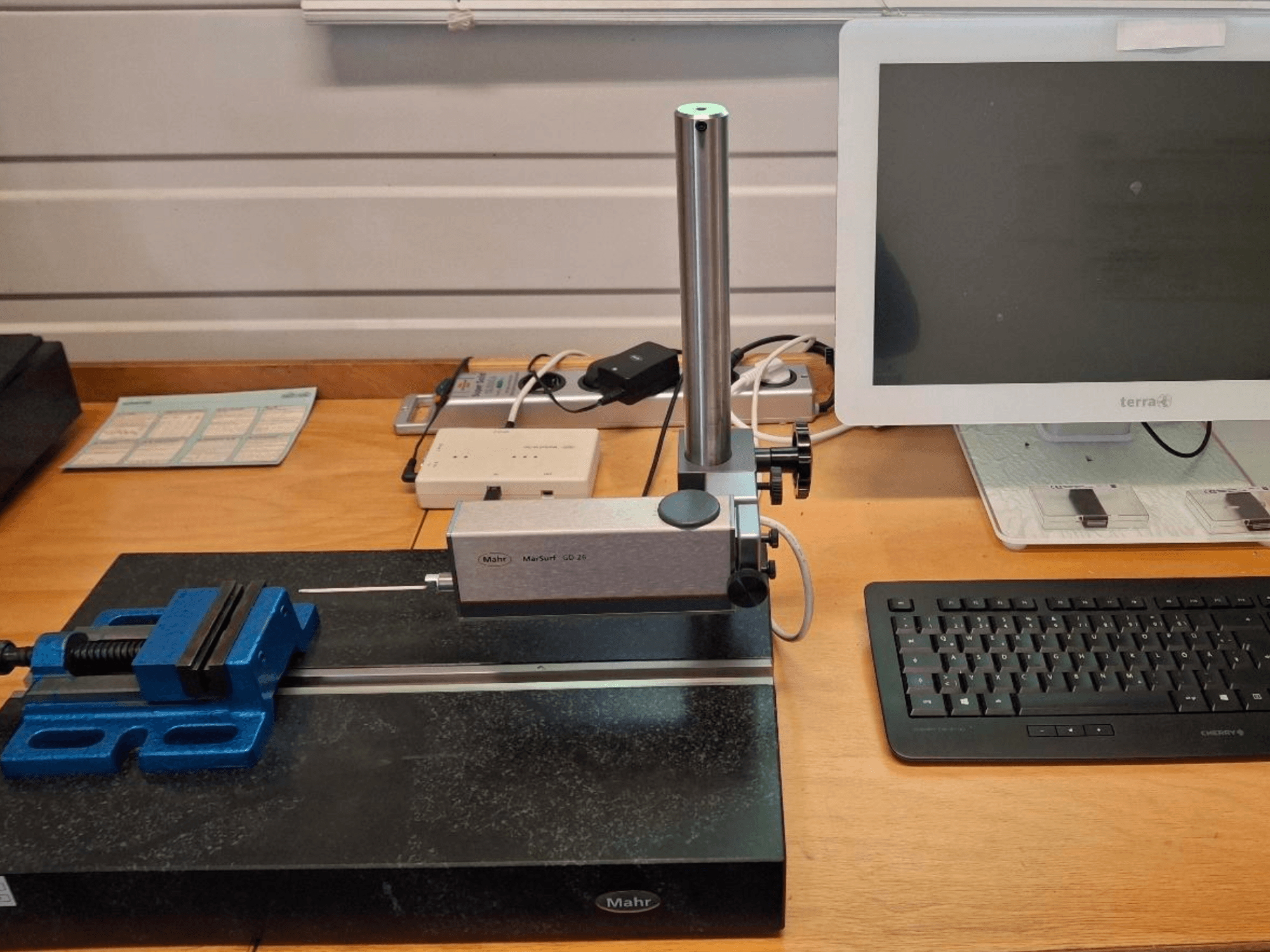 MAHR MARSURF GD 26 CMM op een werkbank, met een meettaster, een blauwe bankschroef en een computermonitor. Vooraanzicht.