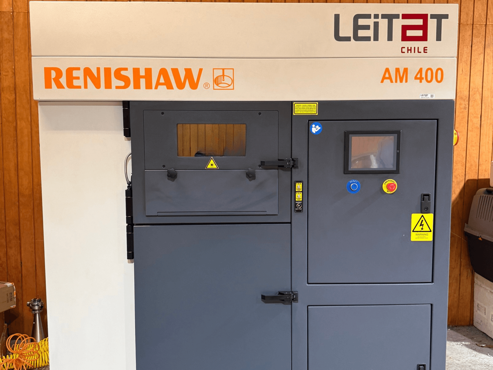 Vooraanzicht van de RENISHAW AM400 3D-printer voor metaal, met bedieningspaneel, waarschuwingsstickers en toegang voor onderhoud.