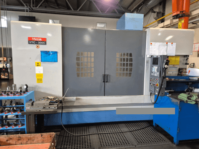 Mazak VTC-300C machine in een fabriek, vooraanzicht met bedieningspaneel, grijze deuren met raamrooster en nabijgelegen apparatuur.