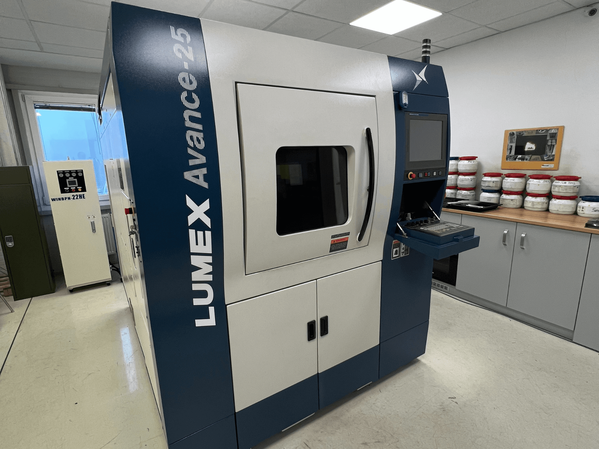 LUMEX Avance-25 3D printer in een lab, vooraanzicht. Bedieningspaneel met scherm en knoppen zichtbaar, apparatuur op achtergrond.