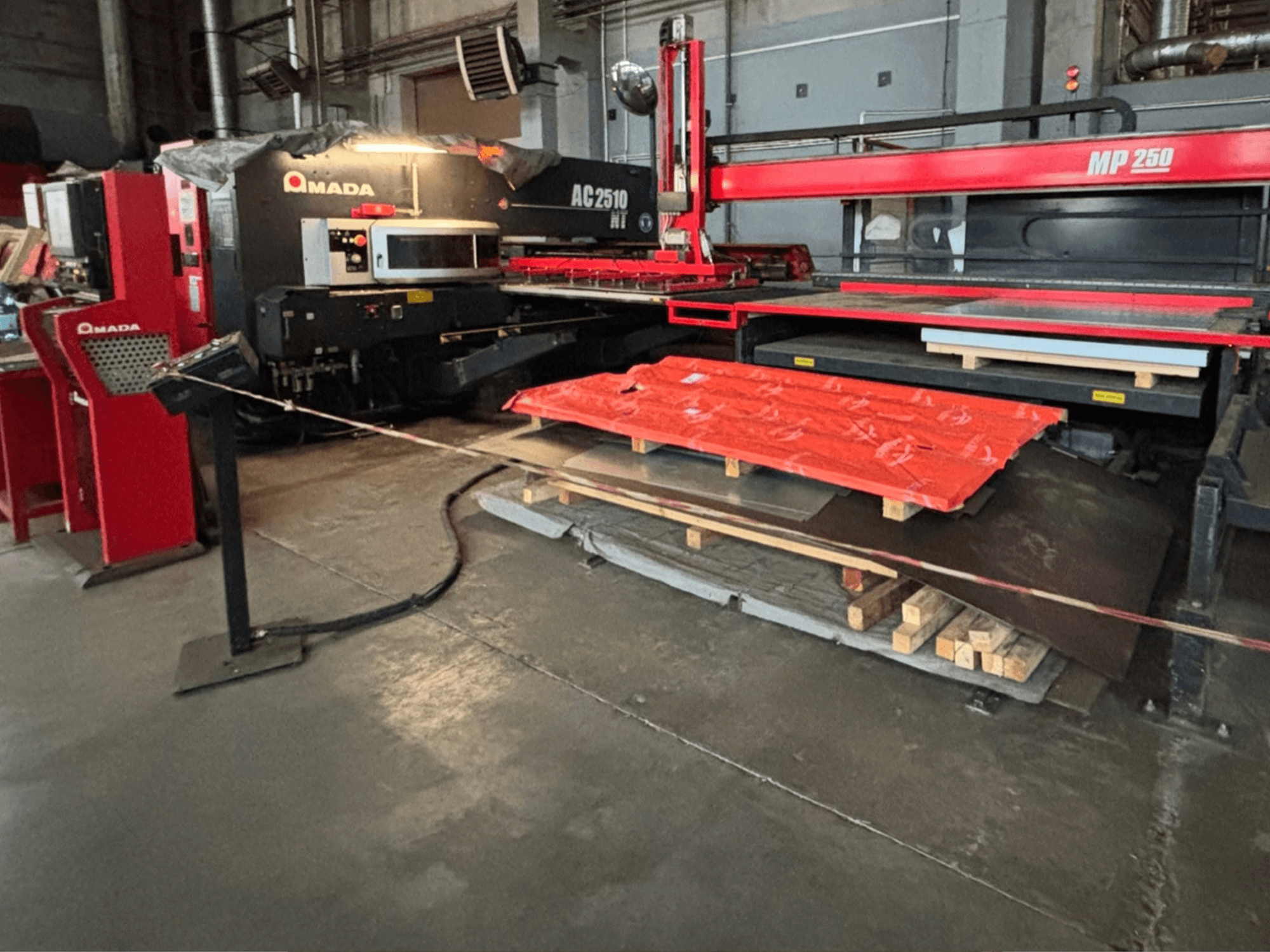 Amada AC2510 NT lasersnijder in fabrieksomgeving, vooraanzicht, zware machines met metalen platen en pallets op de vloer.