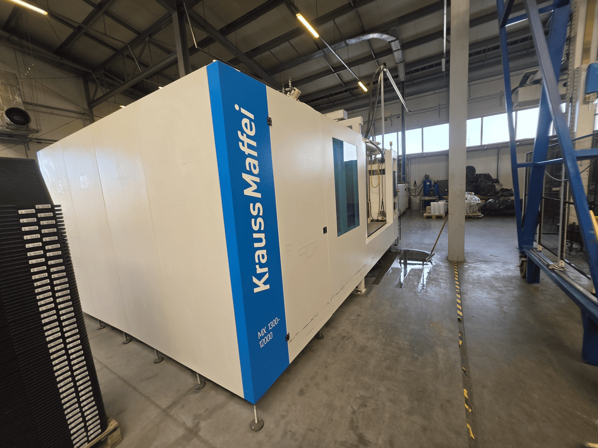 Hydraulische spuitgietmachine Krauss Maffei MX 1300-12000 in zijaanzicht, met het grote witte chassis en de blauwe merklogo’s.