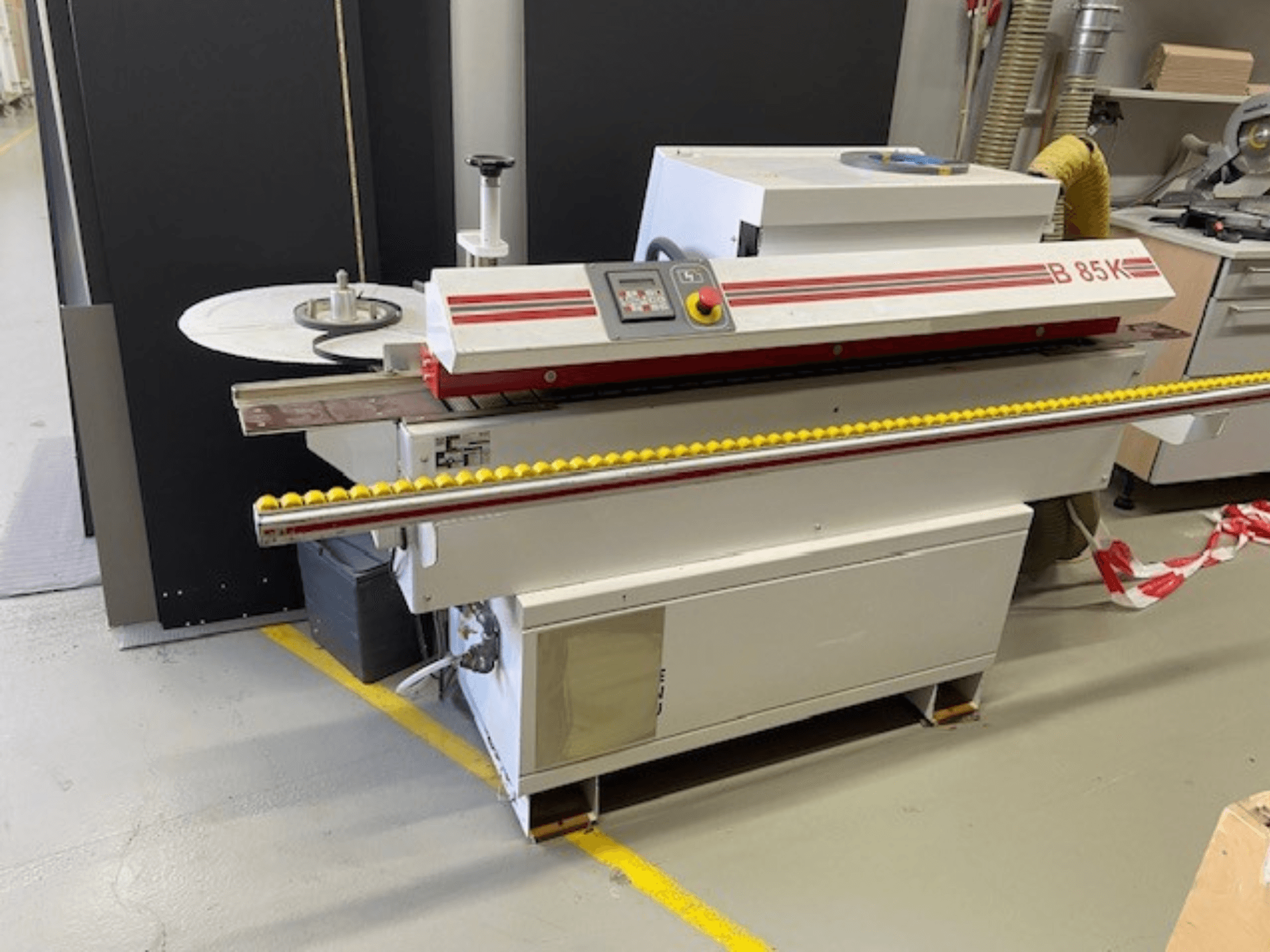 B 85K kantenaanlijmmachine in wit en rood, vooraanzicht, heeft een bedieningspaneel en een gele rol voor de materiaaltoevoer.