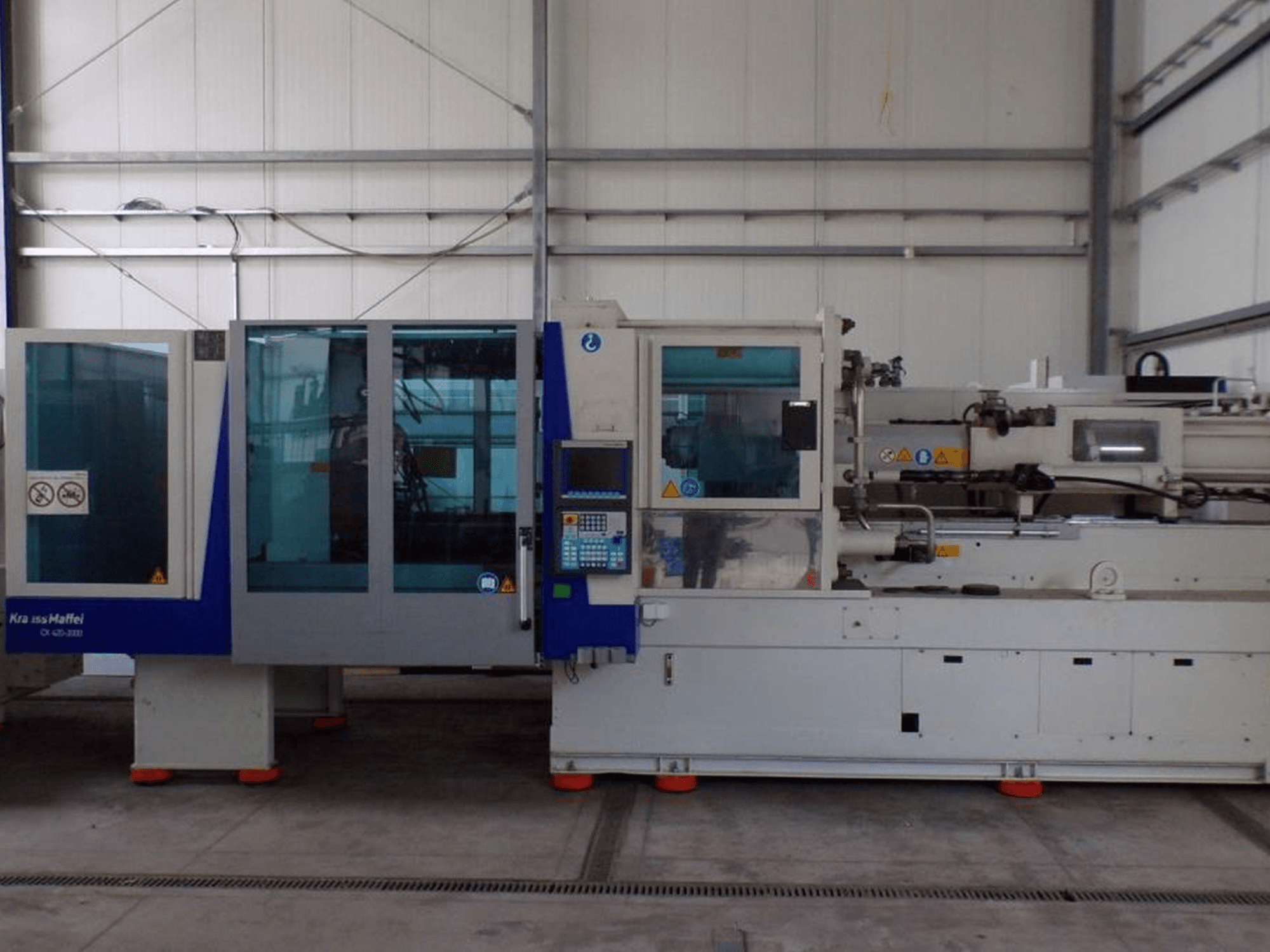 Spuitgietmachine van KraussMaffei, zijaanzicht, met bedieningspaneel en veiligheidsindicatoren, zichtbaar in een fabrieksomgeving.