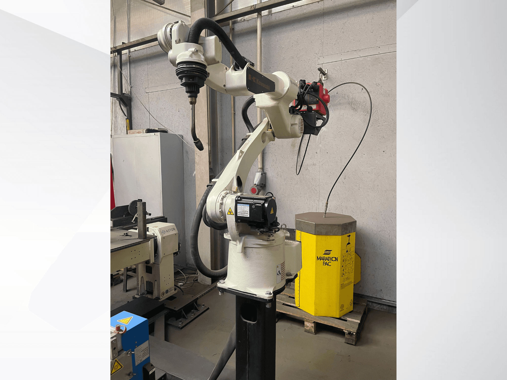 Zijaanzicht van de robotarm van Kawasaki, met een gemonteerde gereedschapshouder en een gele voedingseenheid op de achtergrond.