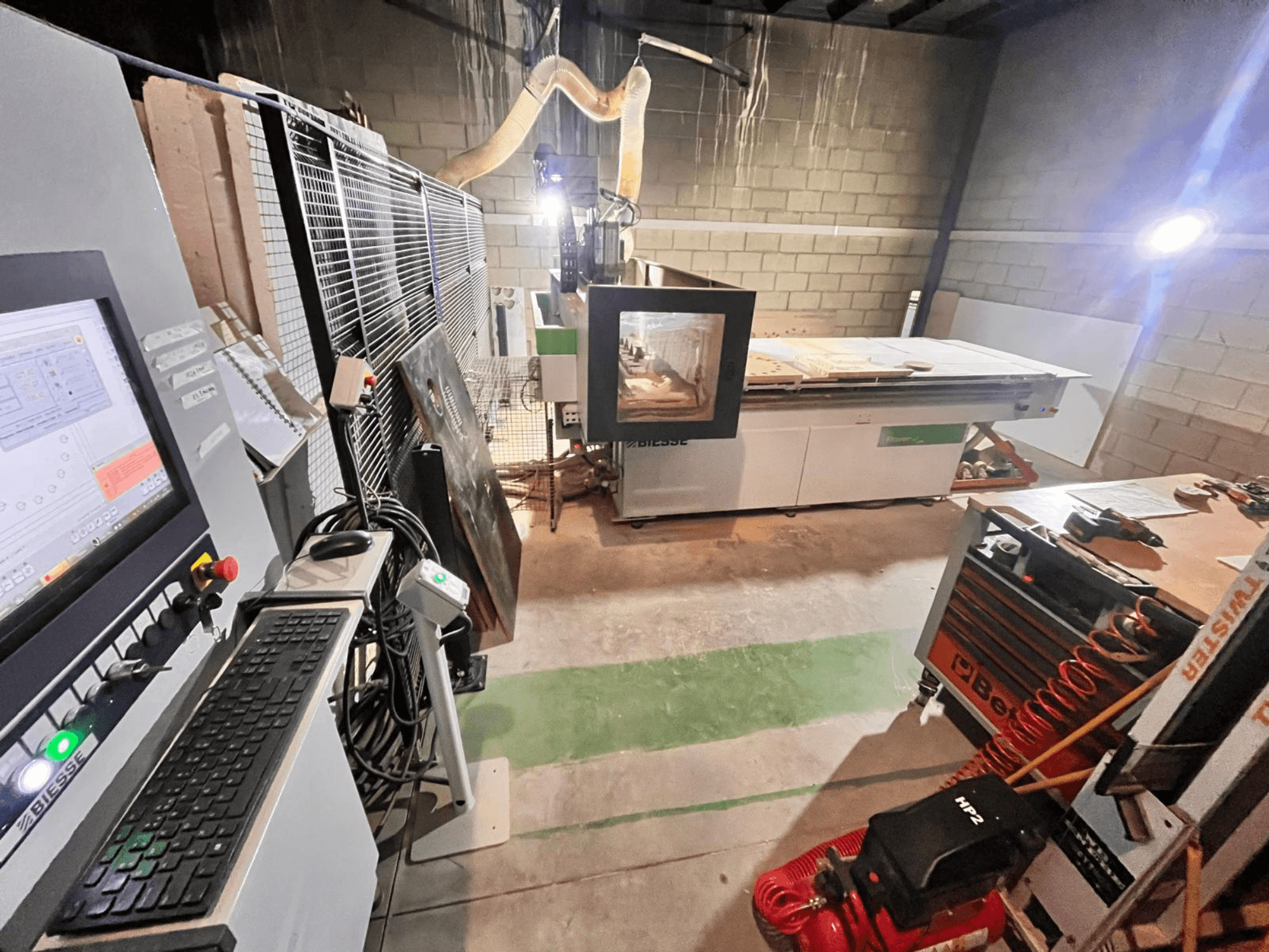 Biesse CNC machine in een werkplaats, zijaanzicht met bedieningspaneel, stofafzuiging, gereedschappen en verschillende materialen op de tafel.