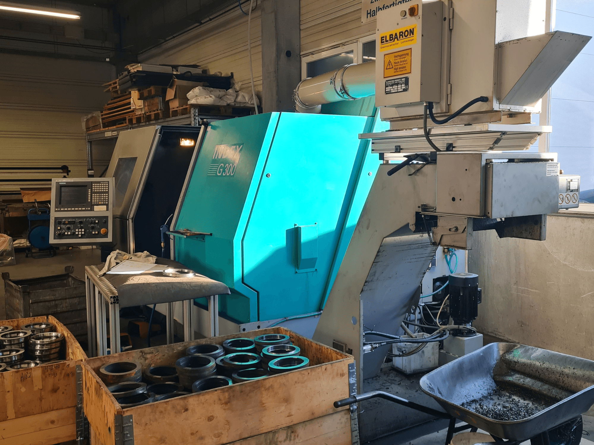 Vooraanzicht van de Index G300 RatioLine horizontale draaimachine met bedieningspaneel, omringd door onderdelen en een transportband.