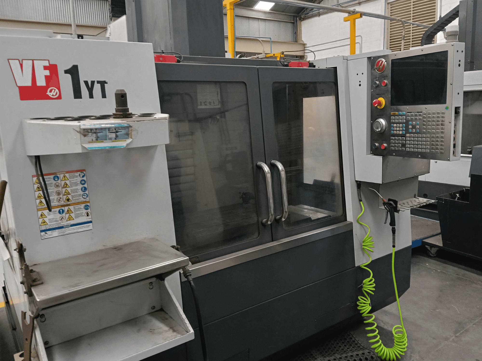 HAAS VF-1YT-2 verticaal bewerkingscentrum, vooraanzicht, met weergave van het bedieningspaneel, de gereedschapshouder, de veiligheidslabels en het bewerkingsgebied.