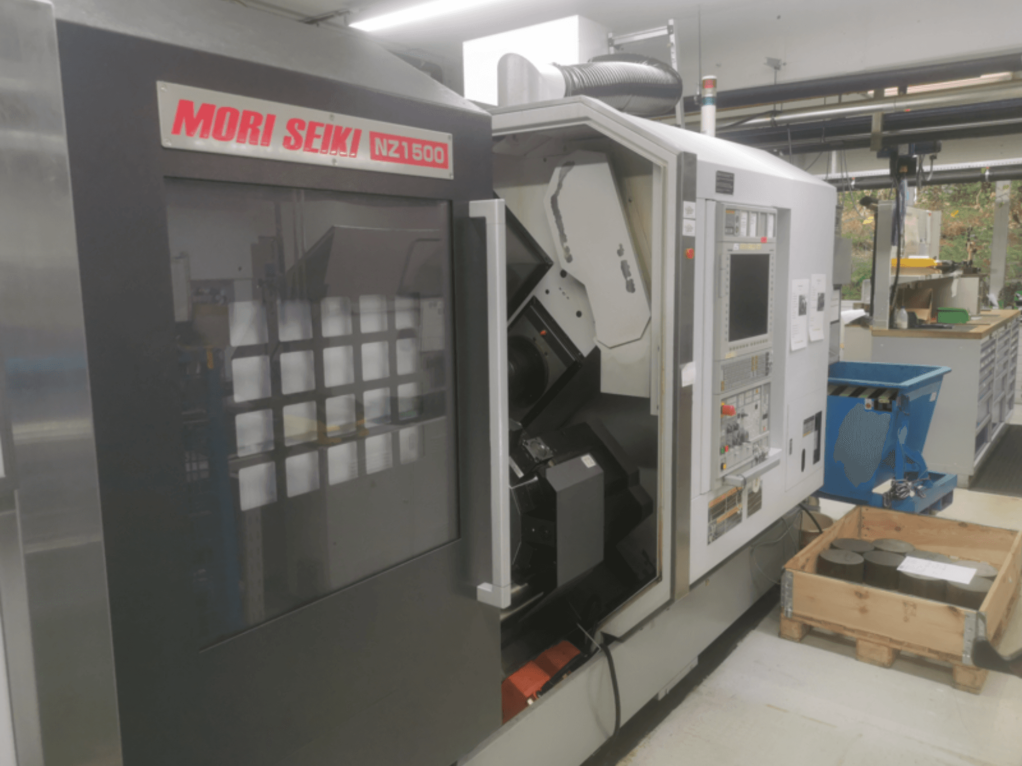 MORI SEIKI NZ1500 CNC machine onder een lichte hoek, met bedieningspaneel en werkruimte met metalen werkstukken op een houten pallet.