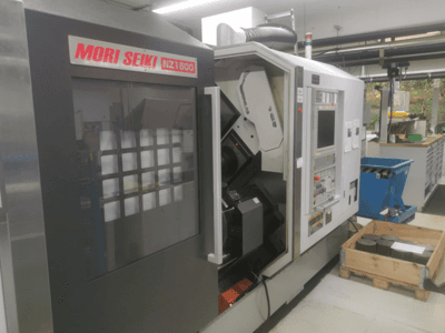 MORI SEIKI NZ1500 CNC machine onder een lichte hoek, met bedieningspaneel en werkruimte met metalen werkstukken op een houten pallet.