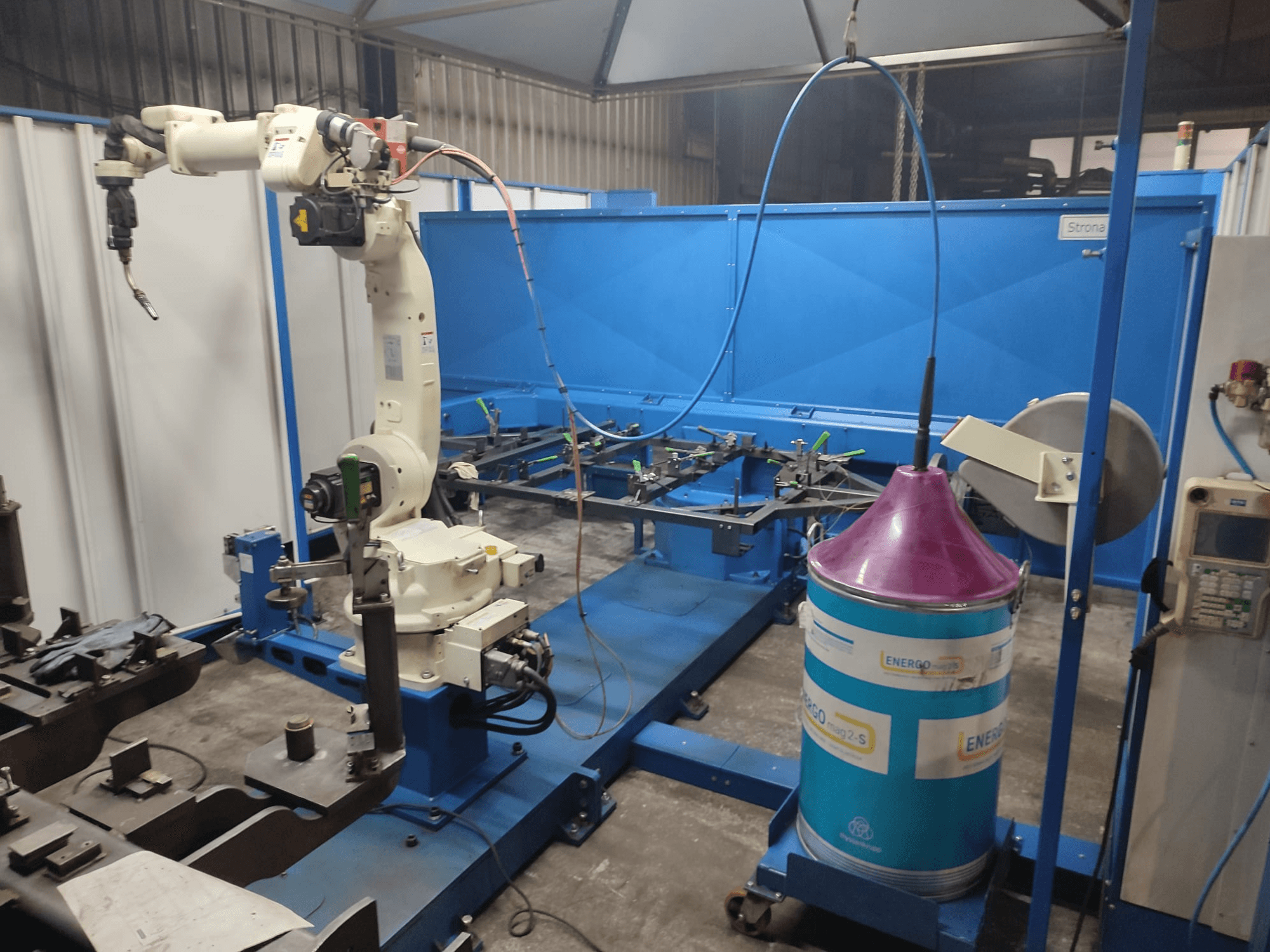 Industriële robotarm met een witte en blauwe basis, opgesteld in een werkplaats met een blauw werkoppervlak en metalen onderdelen.