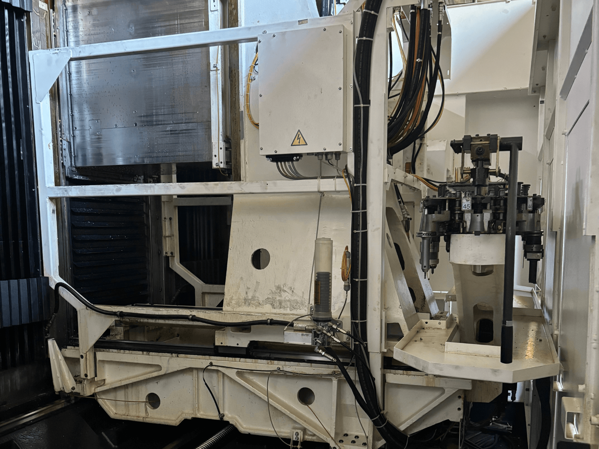 Zijaanzicht van een industriële machine met een witte CNC freesmachine met zichtbare gereedschappen, leidingen en bedieningspaneel.