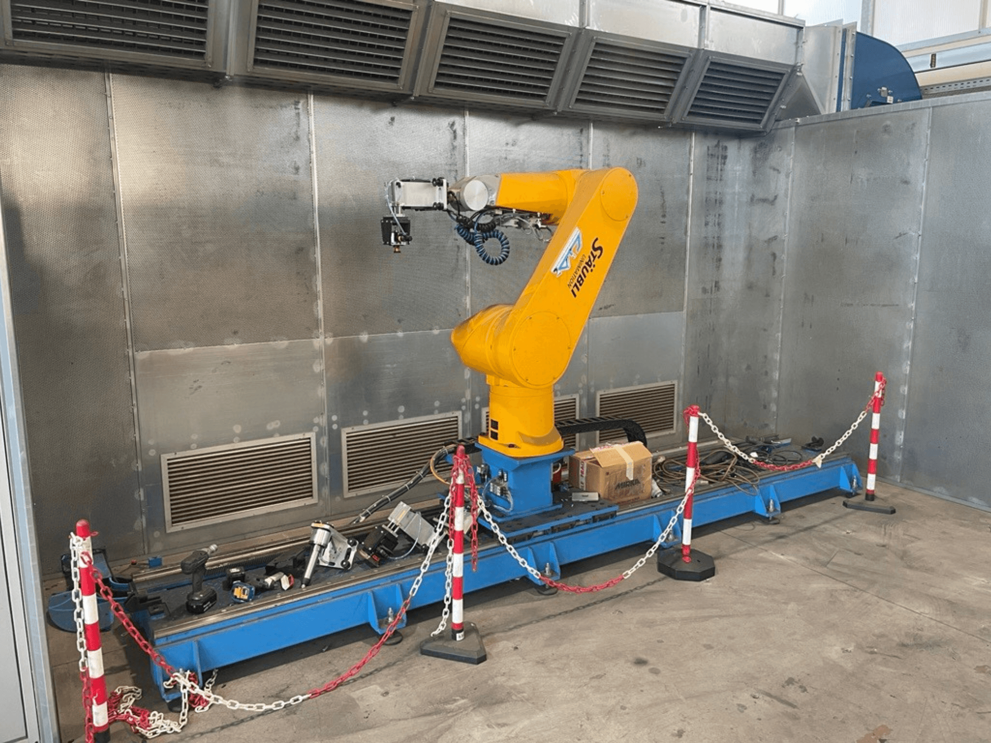 STAUBLI CS8C-RX160 robotarm in een werkplaats, zijaanzicht, met kabels en diverse gereedschappen op een blauwe basis omgeven door beschermende barrières.