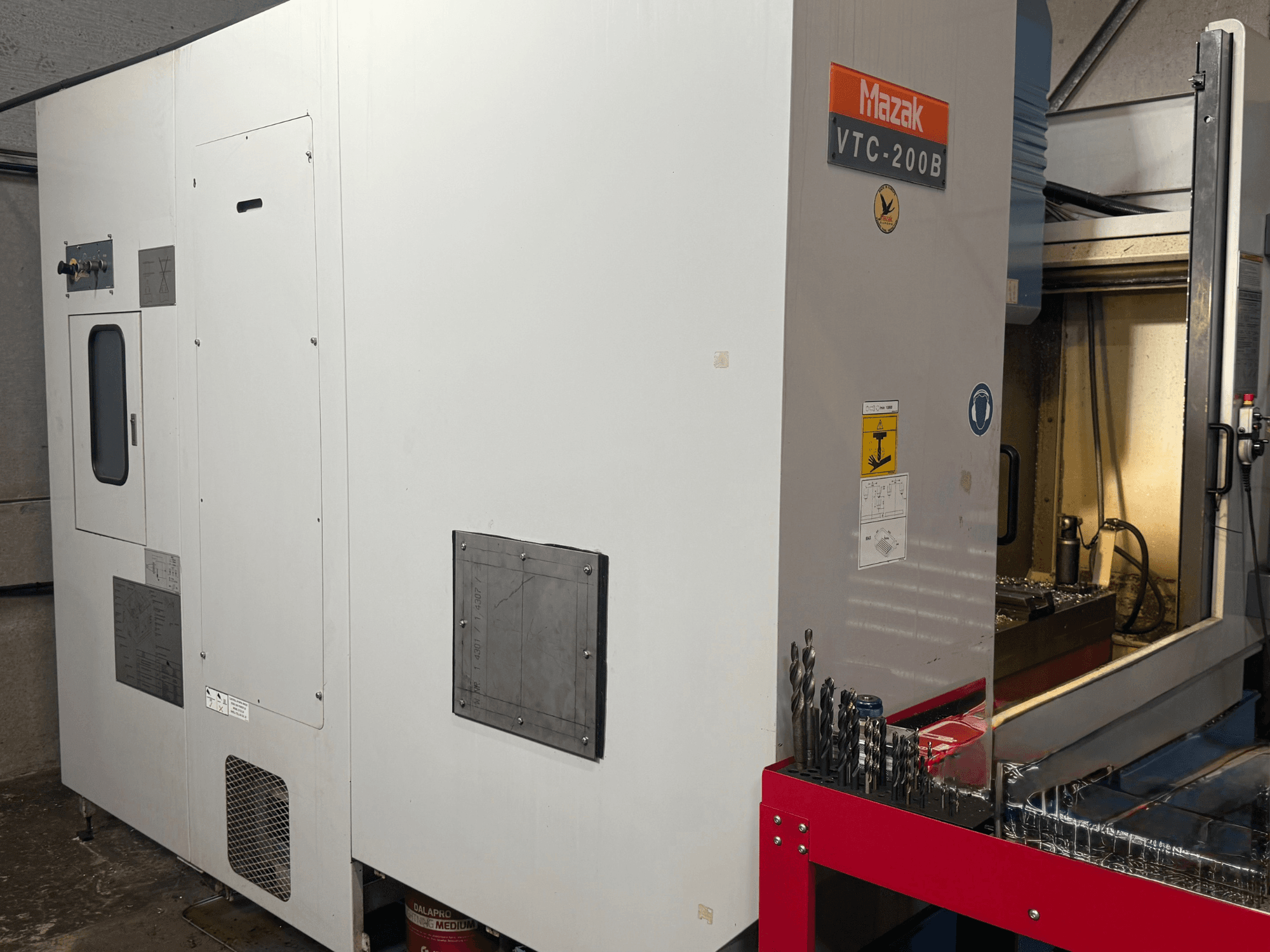 Verticaal bewerkingscentrum Mazak VTC-200B in zijaanzicht, met gelabelde bedieningselementen en boorbits op een rood opbergrek.