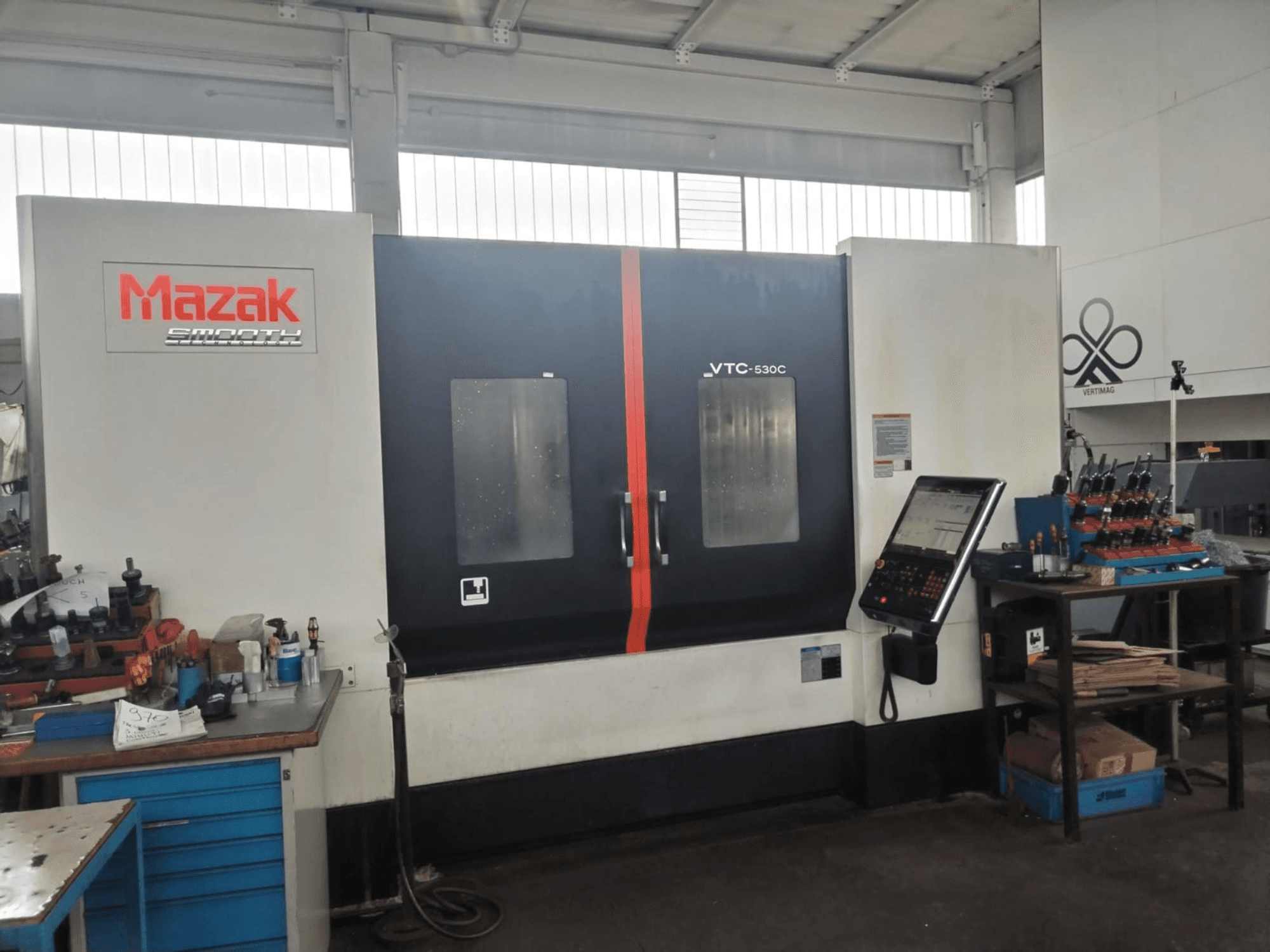 Een verticaal bewerkingscentrum van het type Mazak VTC-530C in een werkplaats, met een bedieningspaneel en gereedschap op een zijtafel.