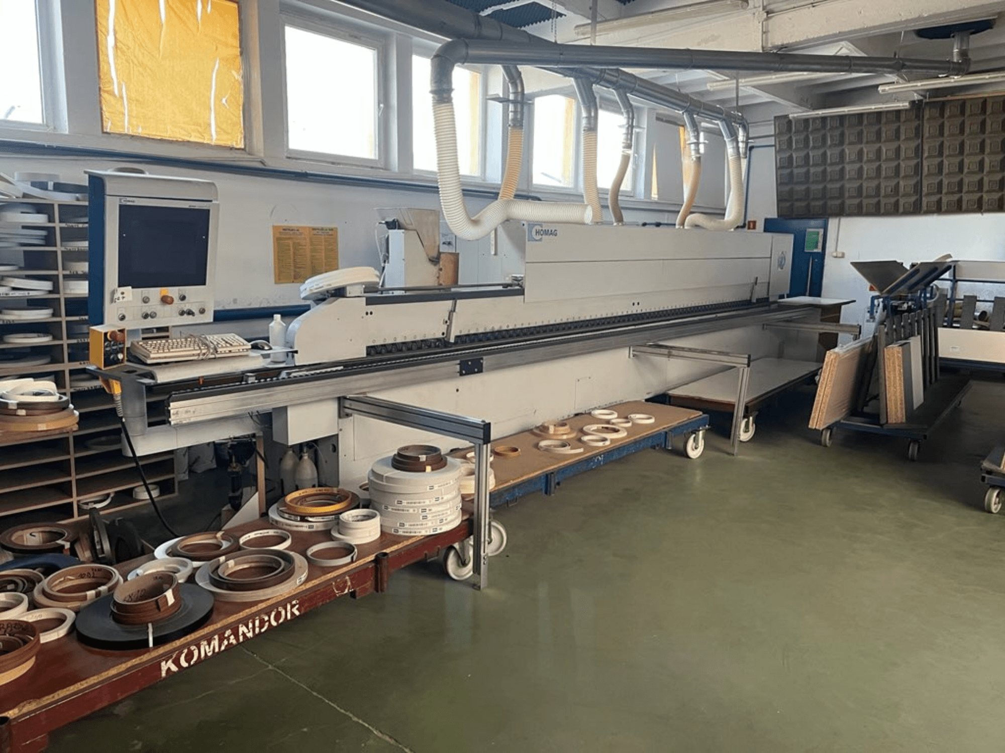 HOMAG kantenaanlijmmachine in een werkplaats, met bedieningselementen en verschillende kantenaanlijmmaterialen op een blauwe kar. Zijaanzicht.