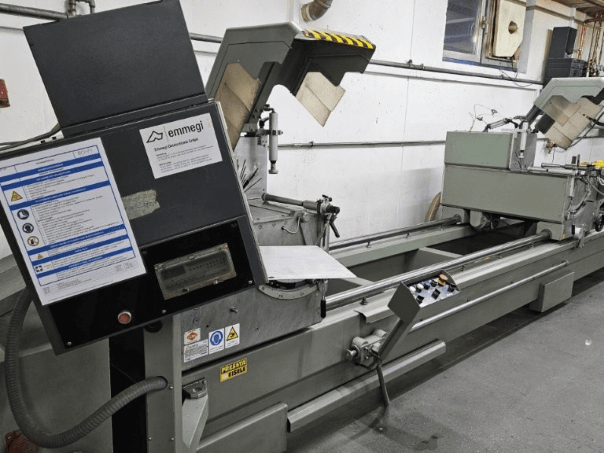 Emmegi industriële snijmachine, vooraanzicht, met bedieningspaneel, veiligheidslabels en operationele onderdelen.