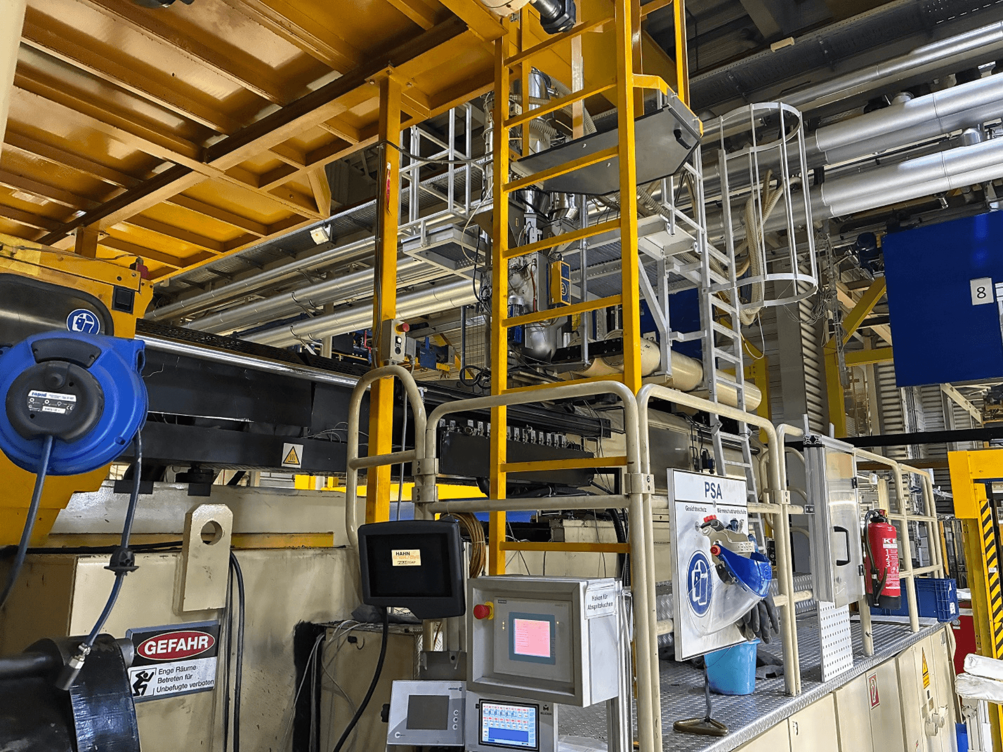 Bedieningspaneel voor industriële machines met blauwe veiligheidsuitrusting en gele steiger, zijaanzicht met pijpen en waarschuwingsborden.