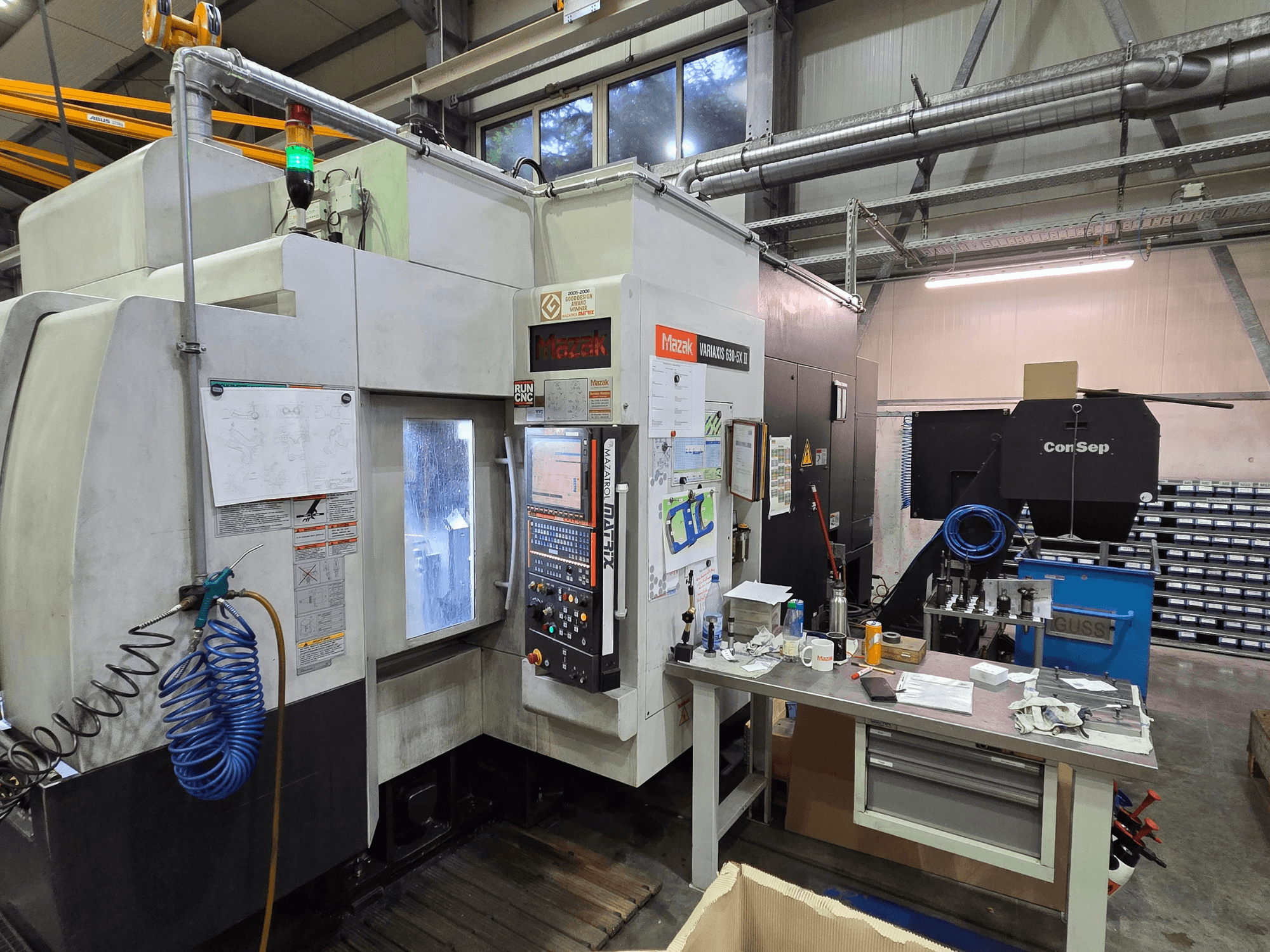 Mazak Variaxis 630-5X CNC machine in een werkplaats, vooraanzicht met bedieningspaneel, slang en accessoires op een zijtafel.