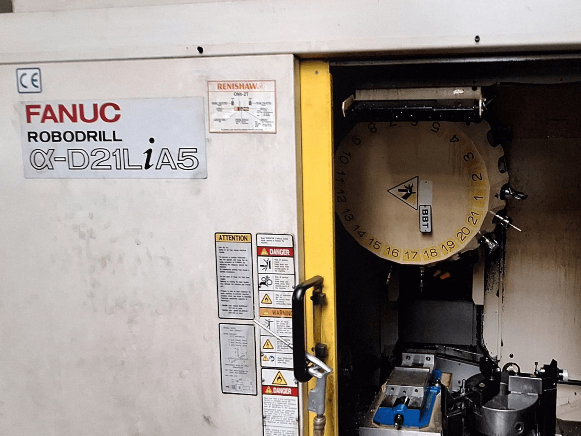 FANUC Robodrill α-D21LiA5, vooraanzicht. Toont gereedschapscarrousel en bewerkingsgebied met klemmen en waarschuwingen op de deur.