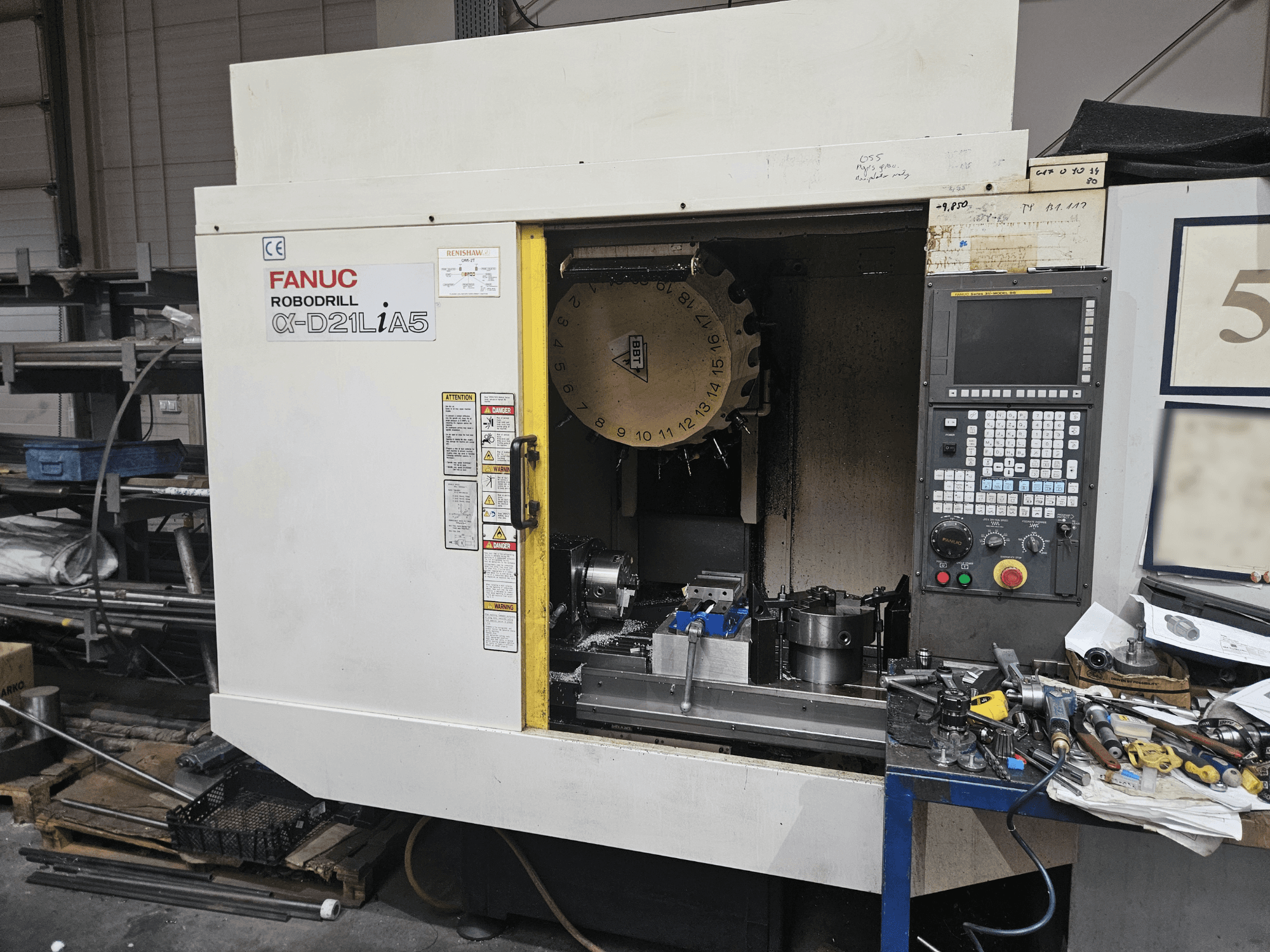 FANUC Robodrill α-D21LiA5, vooraanzicht met gereedschapshouder en bedieningspaneel, omringd door gereedschap en apparatuur in een werkplaats.