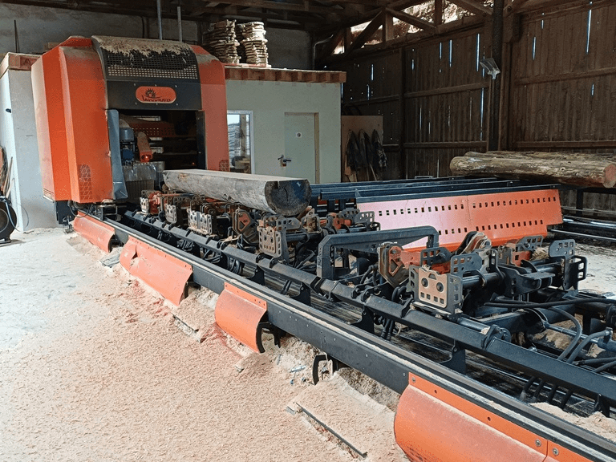 Oranje houtfreesmachine met een boomstam op de transportband, gezien vanaf de voorkant in een werkplaats.