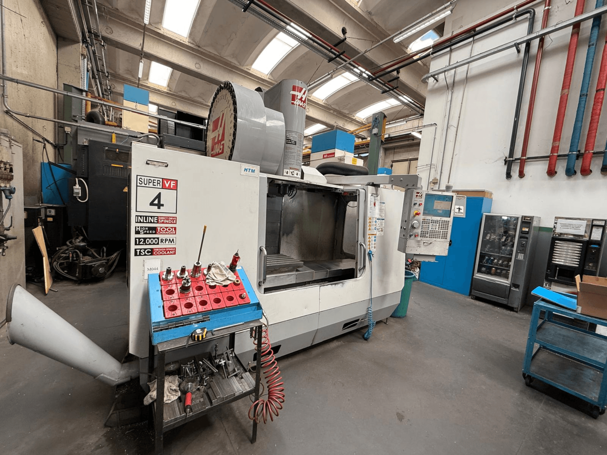 HAAS Super VF 4 CNC freesmachine met een bedieningspaneel, 12.000 tpm spindel en gereedschappen op een blauw opbergrek, zijaanzicht.