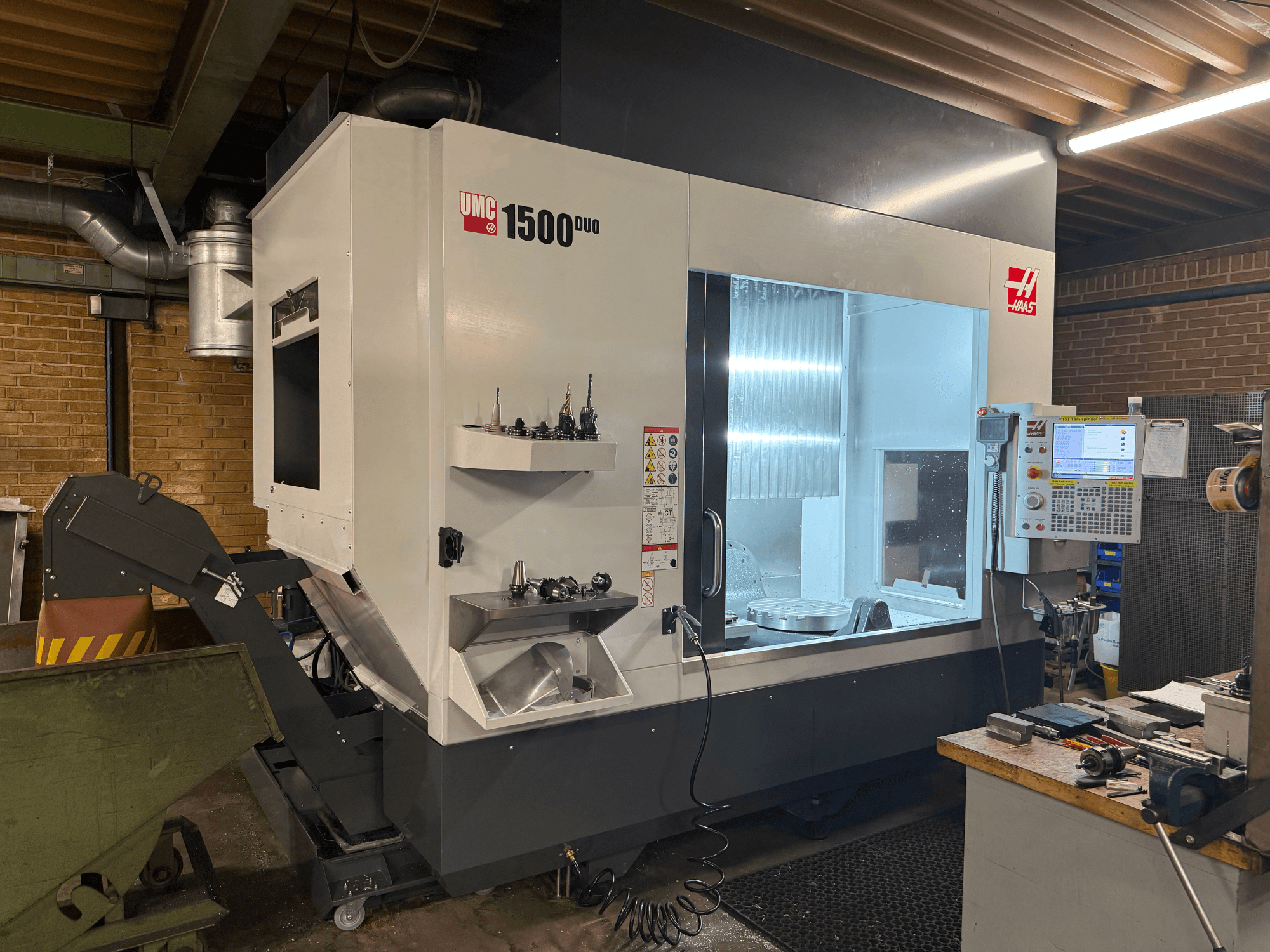 Haas UMC 1500DUO CNC machine, vooraanzicht, met een groot venster, bedieningspaneel en gereedschaphouders op een plank.