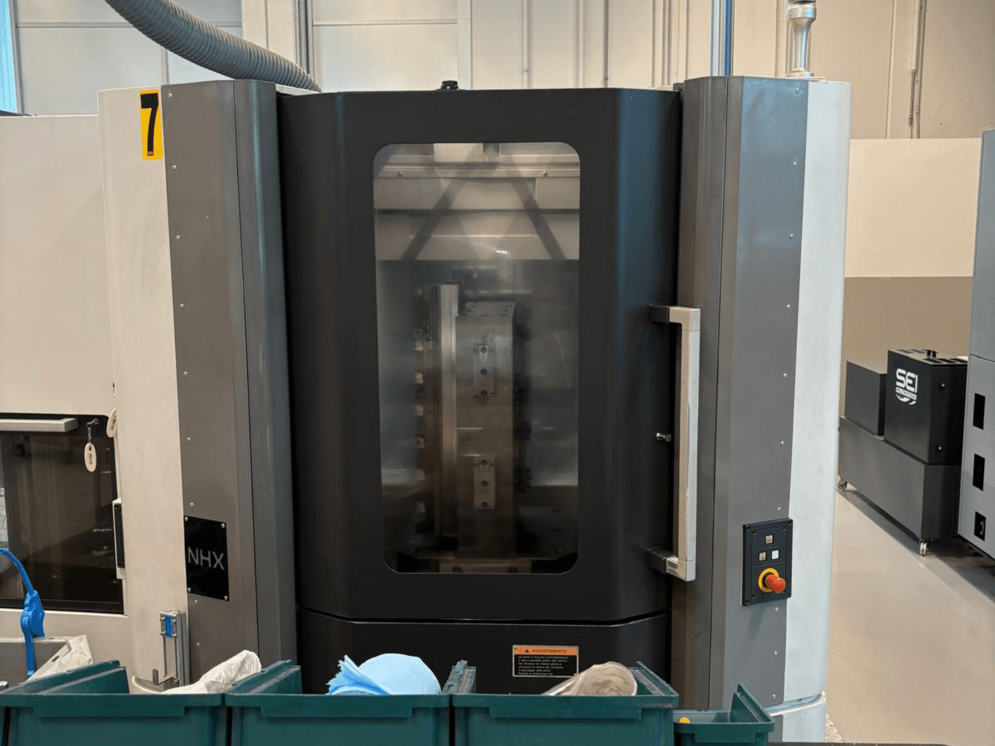 NHX CNC machine in vooraanzicht, met bedieningspaneel en duidelijk display; staat in een werkplaats met groene bakken in de buurt.