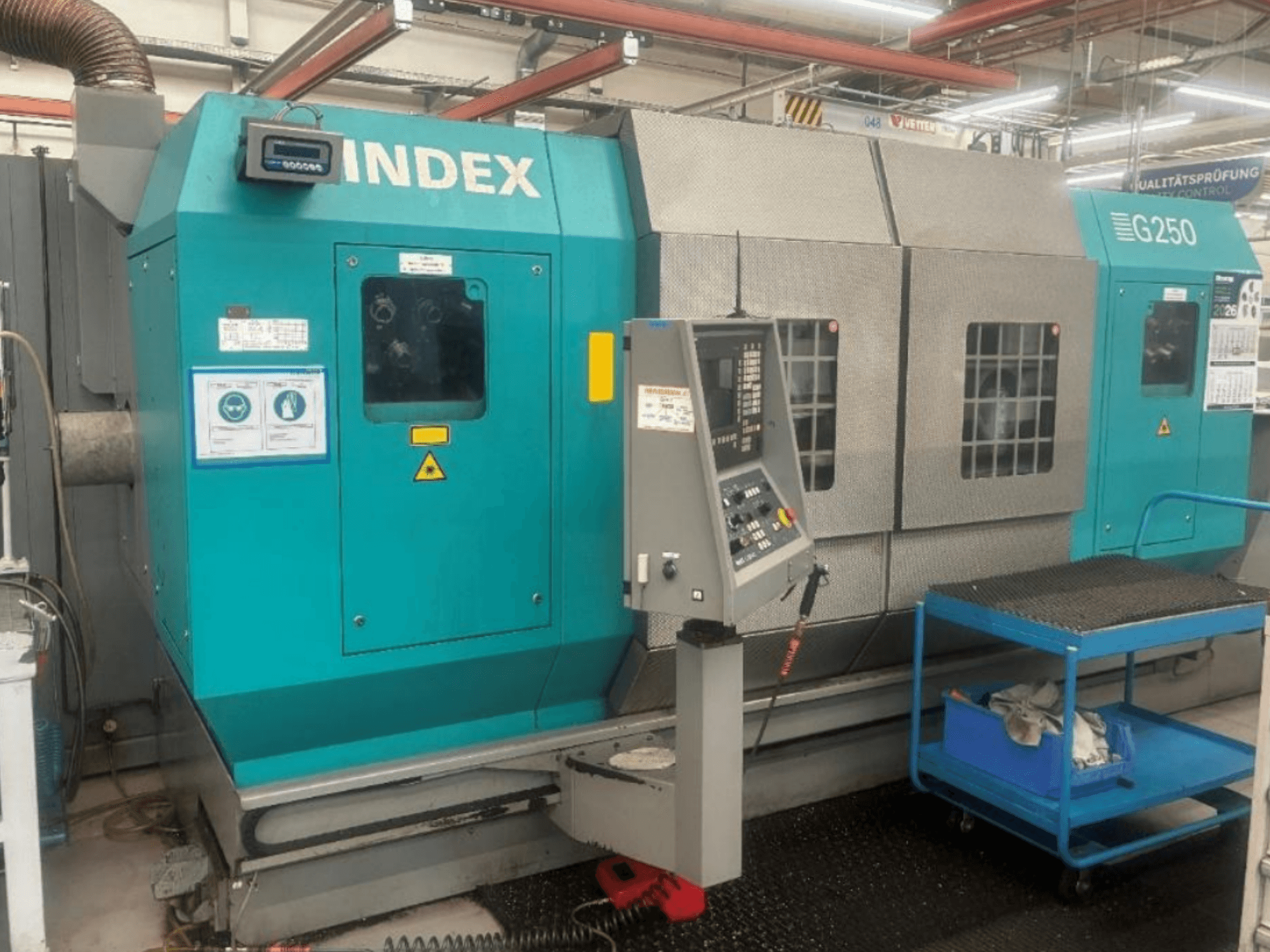 Horizontale draaimachine INDEX G250, vooraanzicht met bedieningspaneel en metalen behuizing, voorzien van veiligheidsstickers en een blauwe kar.