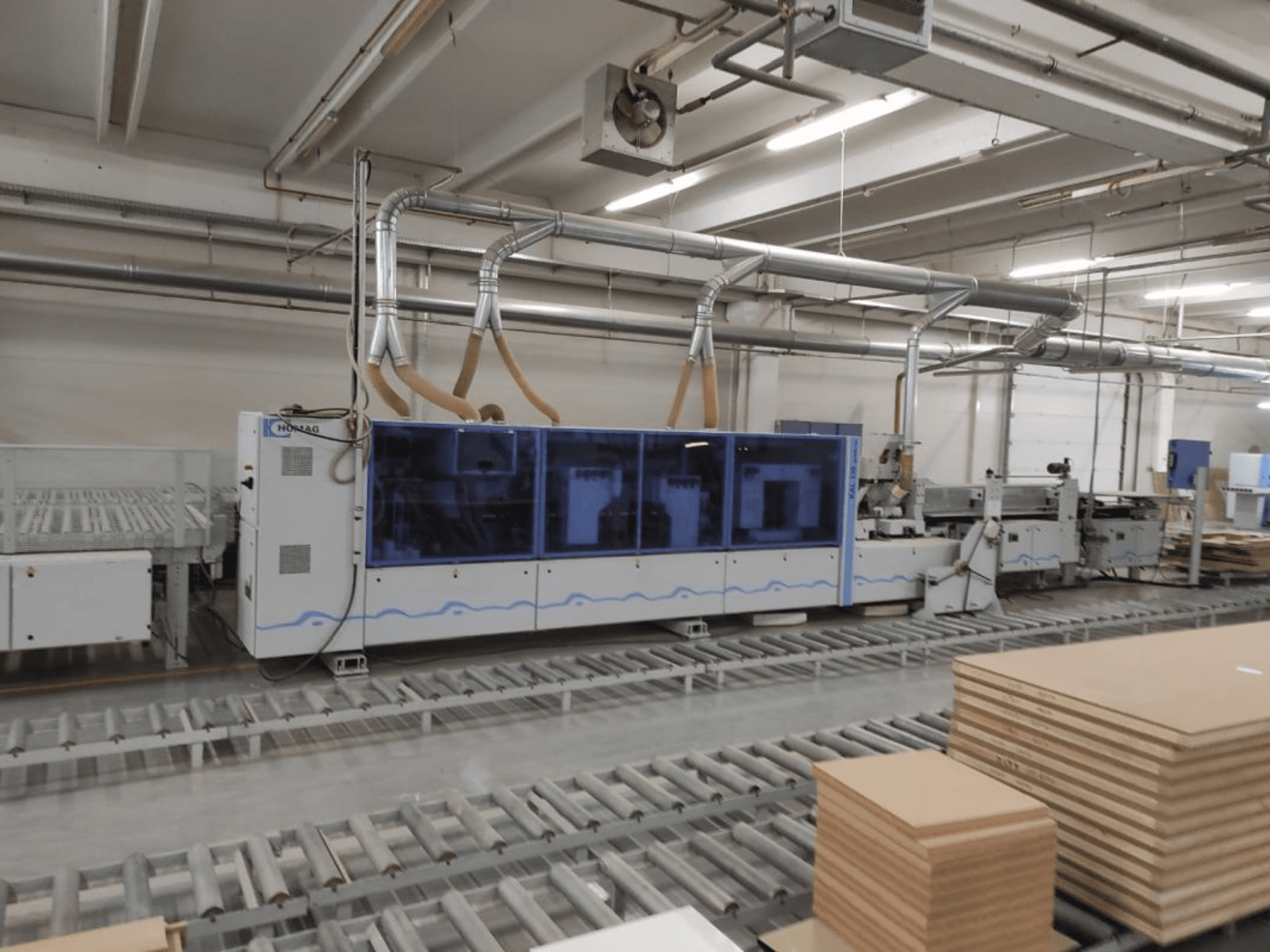 HOMAG productiemachine in een fabriek, vooraanzicht, met blauwe behuizing en transportsysteem met gestapelde multiplex platen.