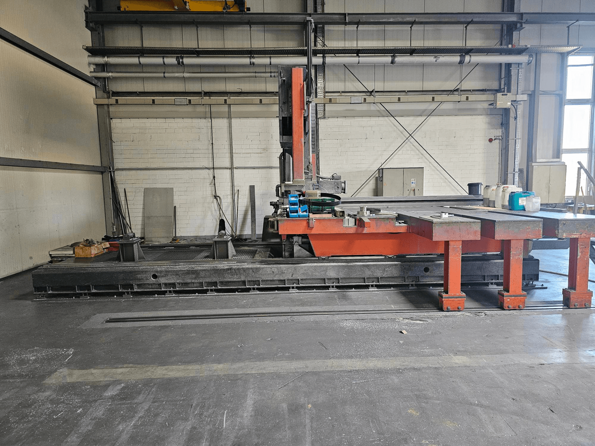 Industriële snijmachine in een open werkplaats, oranje en zwart met een grote tafel; zijaanzicht met accessoires en werkruimte.