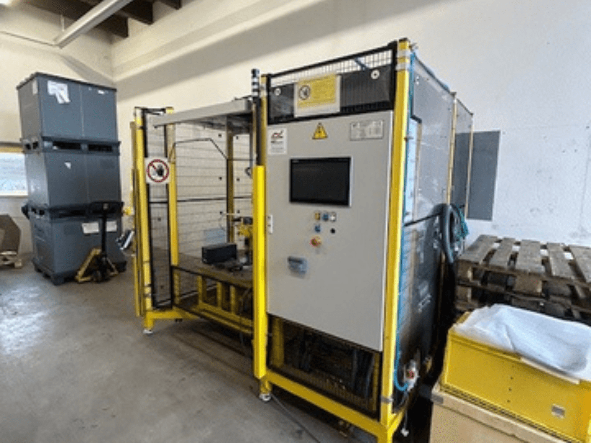 Industriële machine met bedieningspaneel en veiligheidsfuncties, vooraanzicht; geel frame, grijze bakken en pallets op achtergrond.